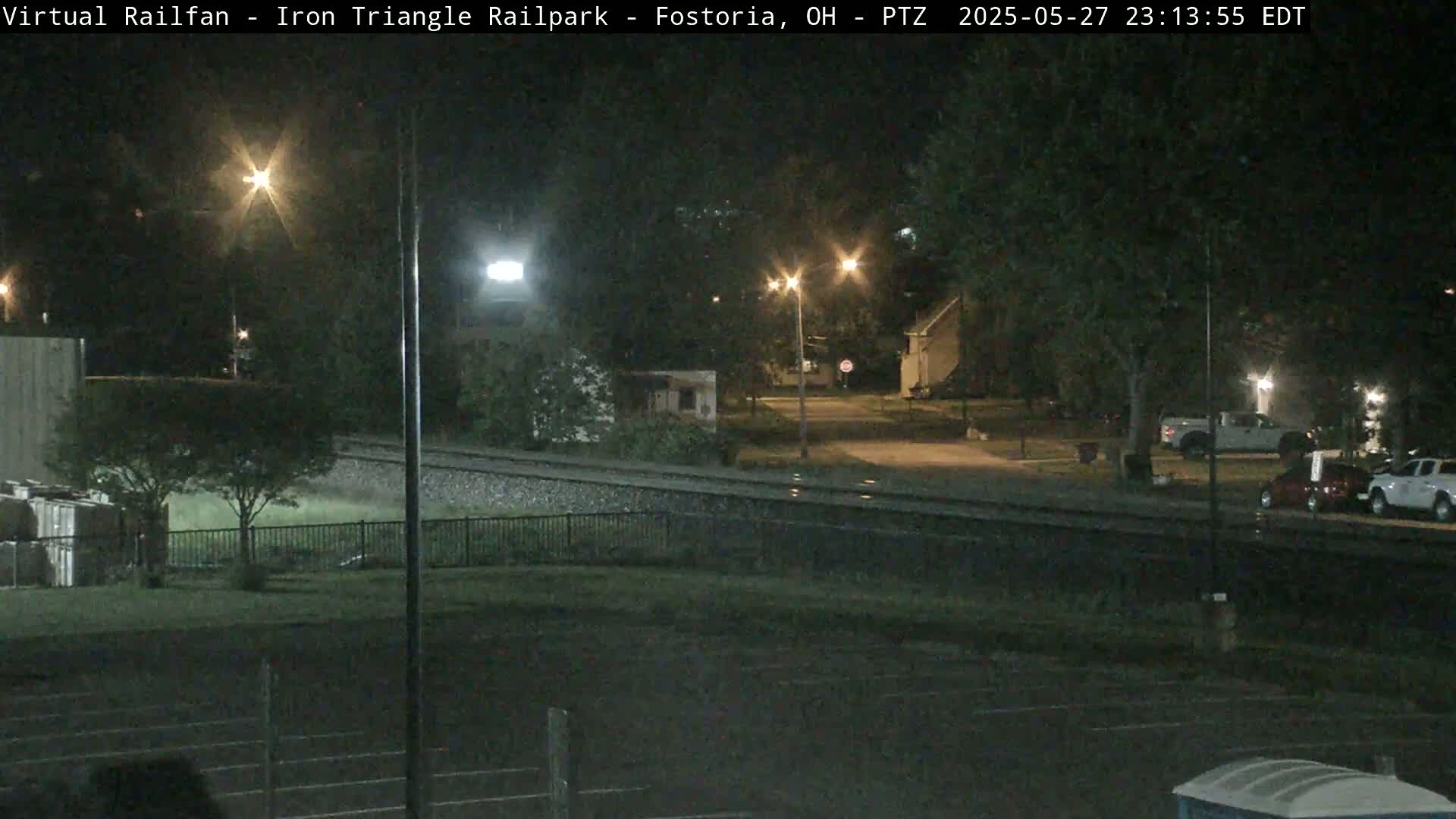 A nighttime view of a rail yard and surrounding area, lit by streetlights, showing parked vehicles and a quiet street.