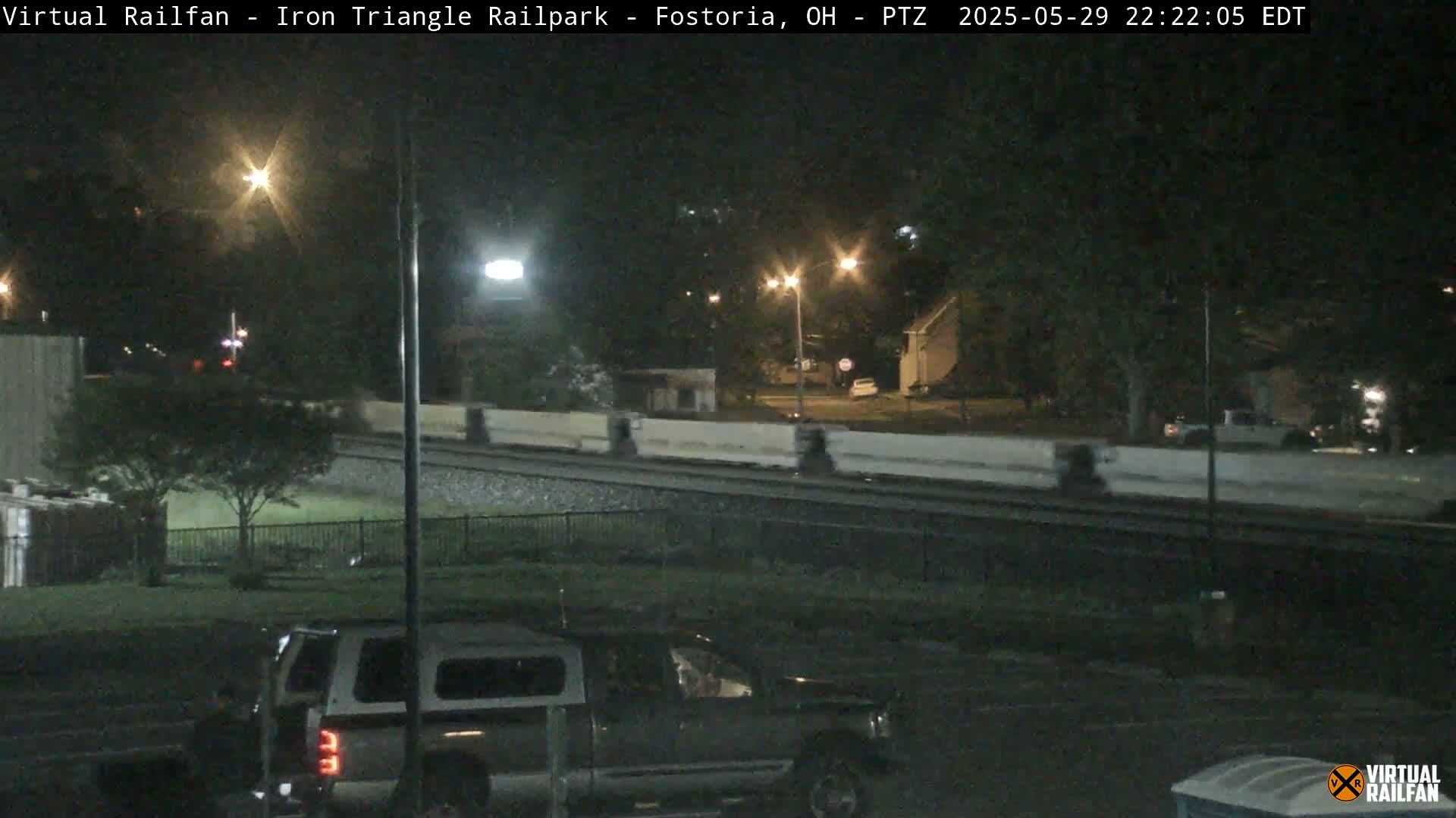 A nighttime view of a train track next to a road with a pickup truck parked nearby, under streetlights.