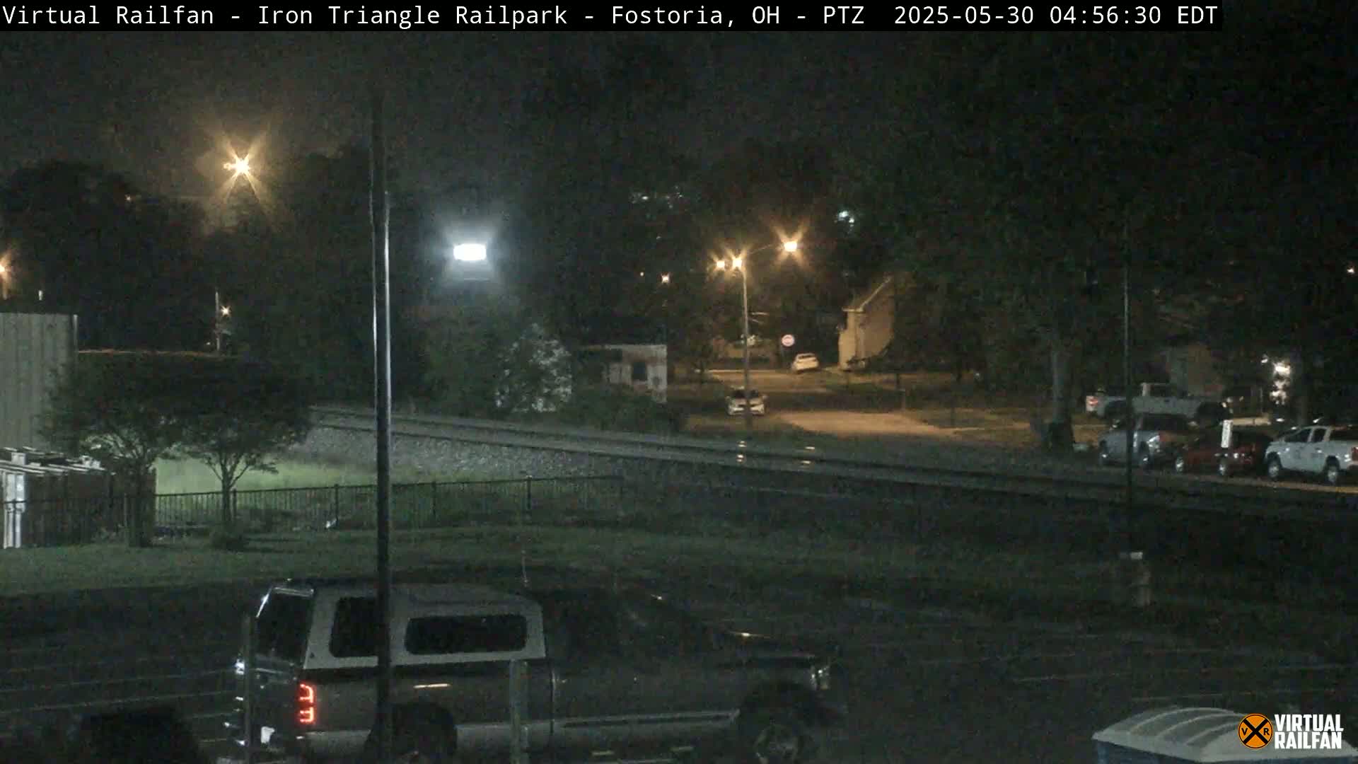 A nighttime view of a railroad crossing near a residential area with several parked vehicles, under dark and wet conditions.