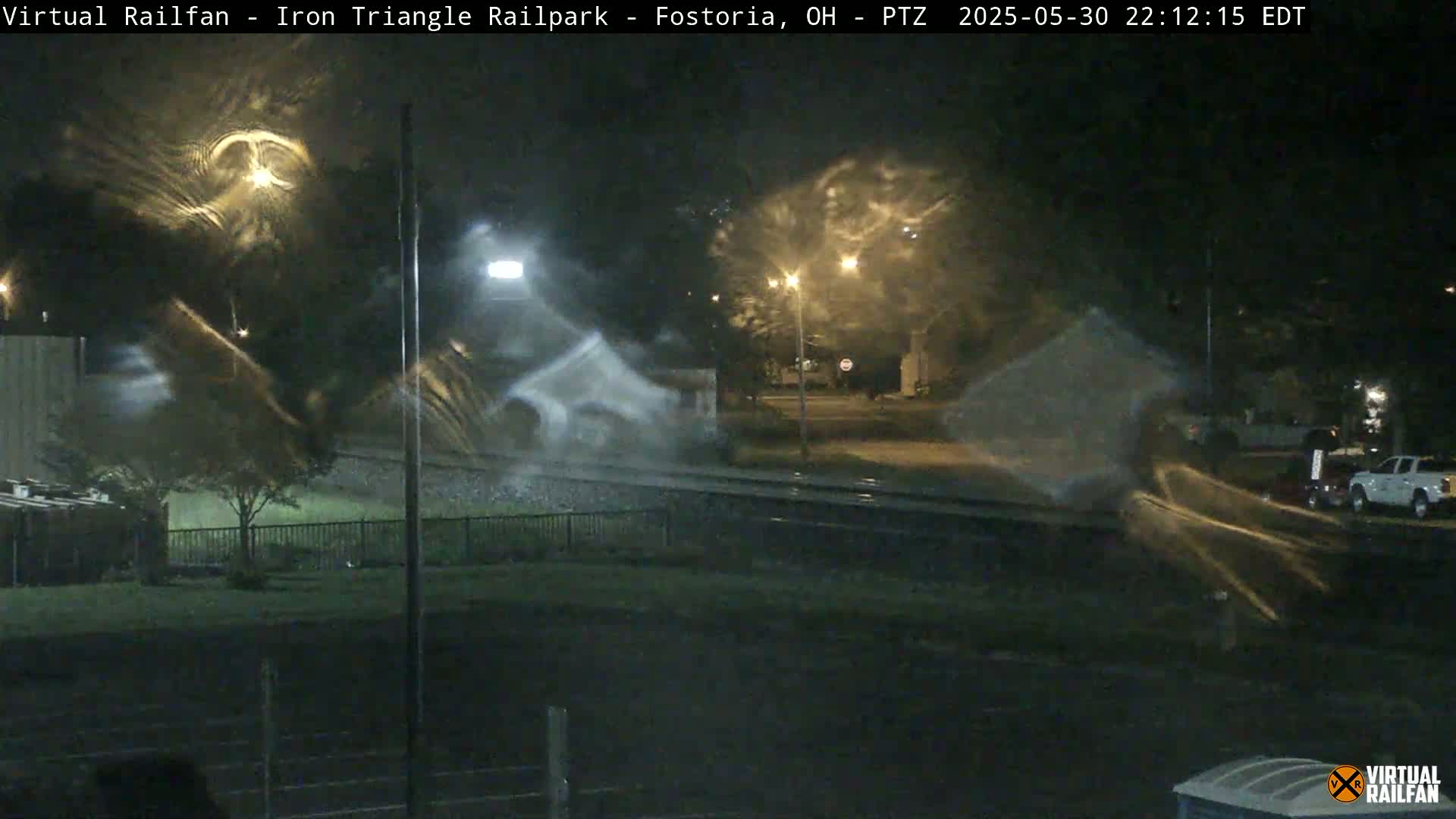 A nighttime, rainy scene shows a rail yard with some parked vehicles and streetlights visible through rain-streaked glass.
