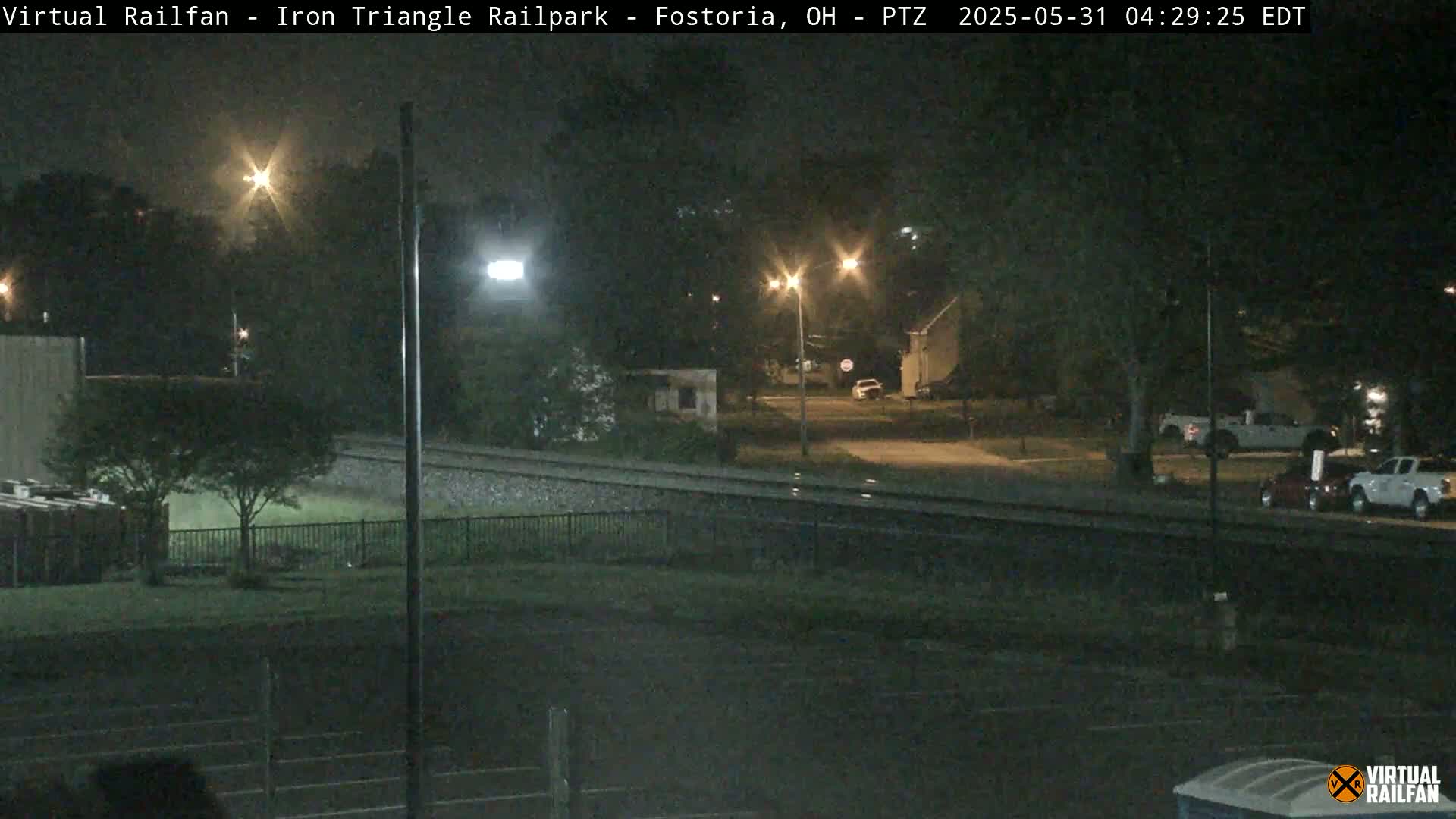 A nighttime view of railroad tracks adjacent to a residential area with streetlights illuminating parked vehicles and surrounding trees under dark, clear skies.