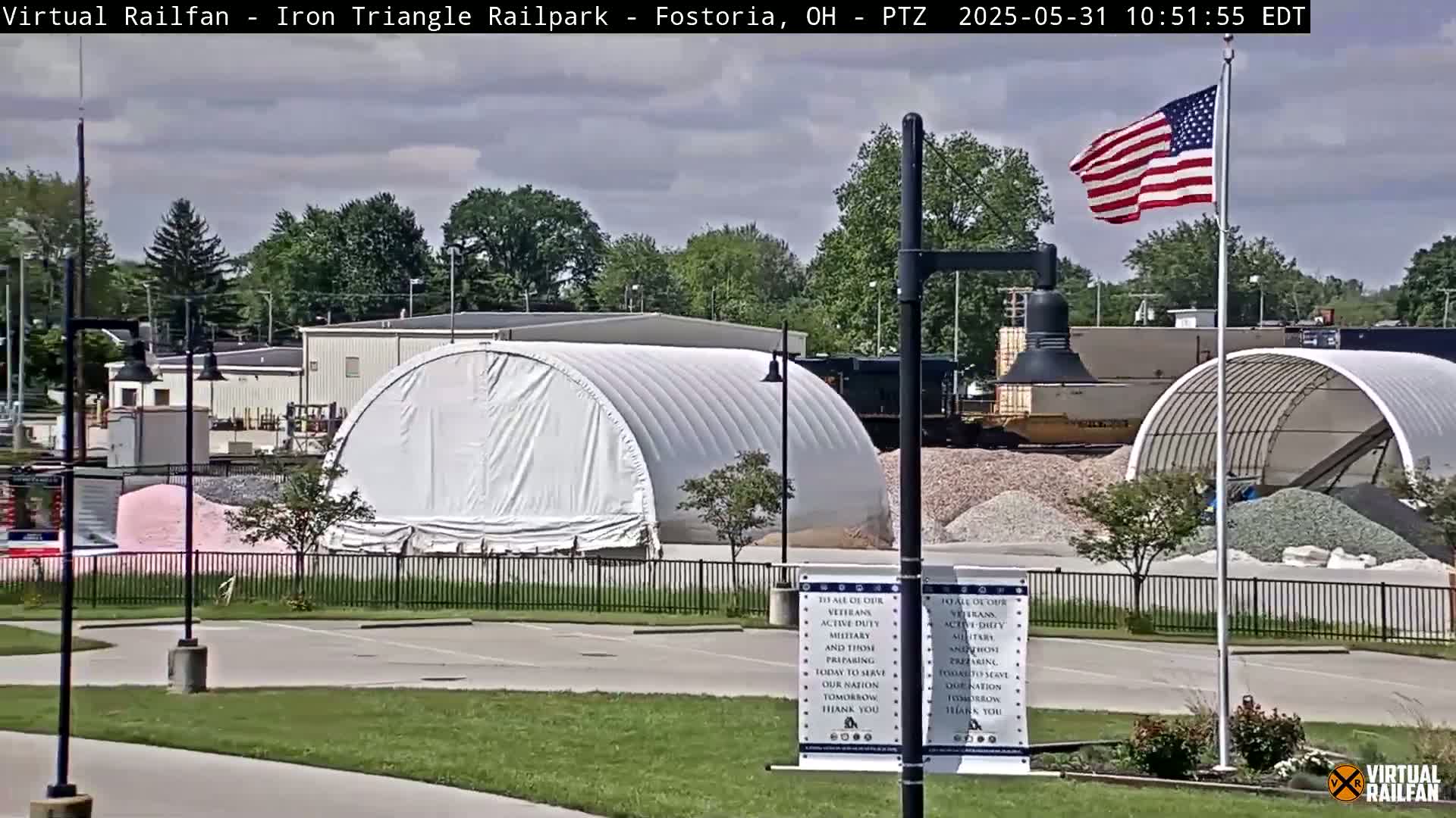 A partly cloudy day reveals an outdoor scene of industrial buildings, piles of aggregate materials, and two large white fabric structures, with an American flag waving in the breeze.