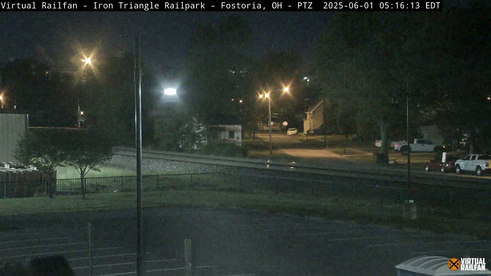 A nighttime view of a railroad crossing near a residential area, with several vehicles parked nearby under partly cloudy skies.