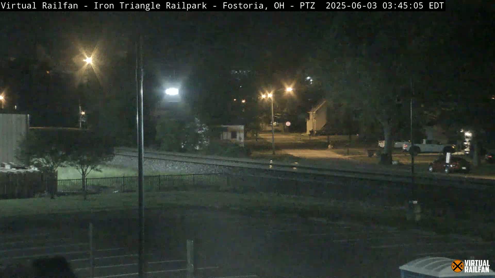 A nighttime view of a railroad track, a road, and a residential area, illuminated by streetlights.