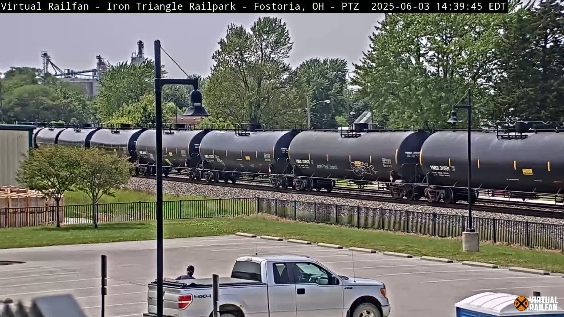 A long freight train of black tanker cars passes by a parking lot where a silver pickup truck is parked, under a partly cloudy sky.
