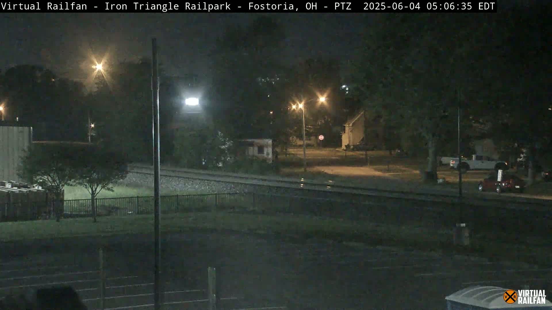 A nighttime view of a railroad track, adjacent to a residential area with streetlights, under slightly wet conditions.