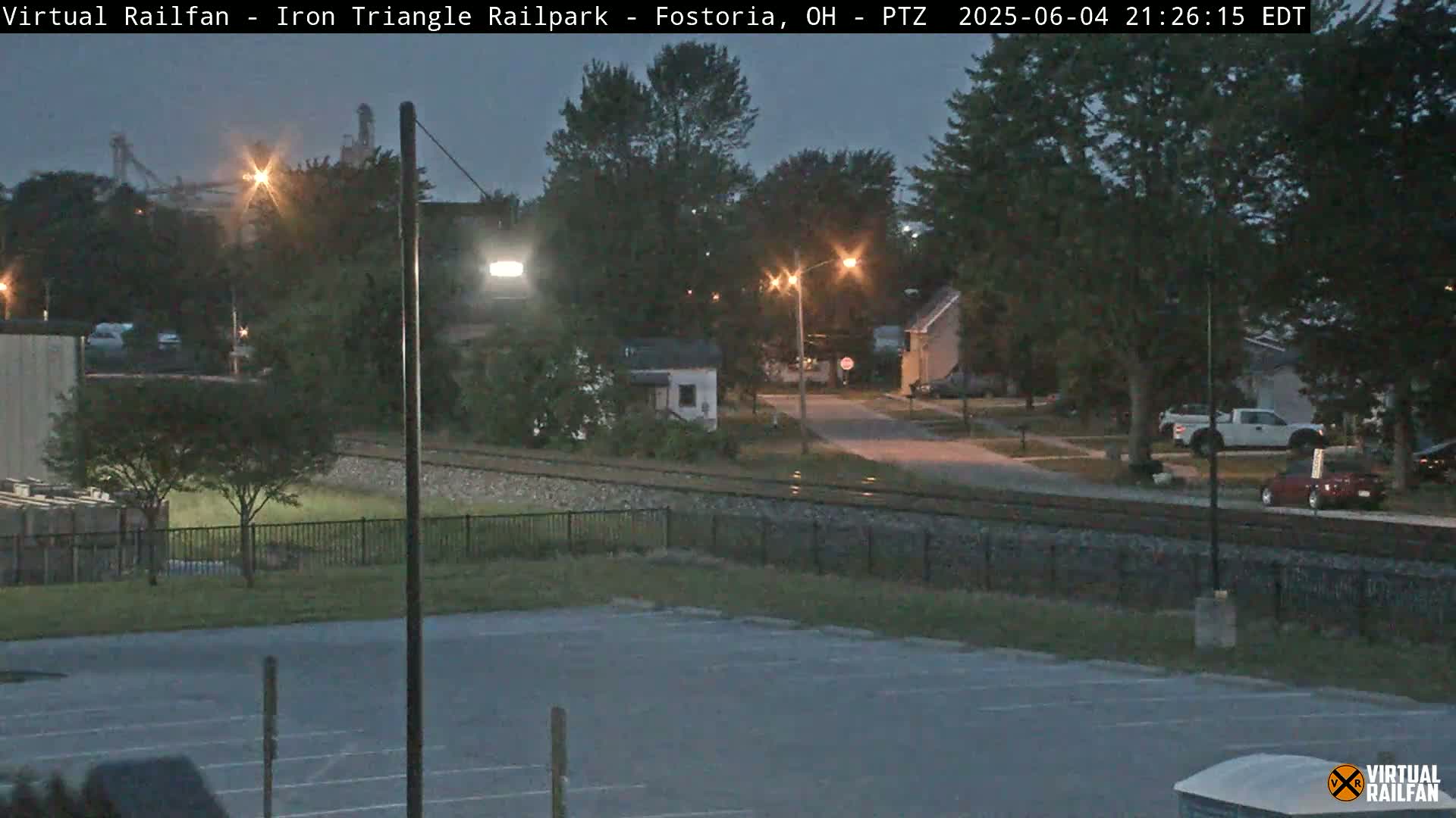 A nighttime view of a residential street near railroad tracks, under a twilight sky.