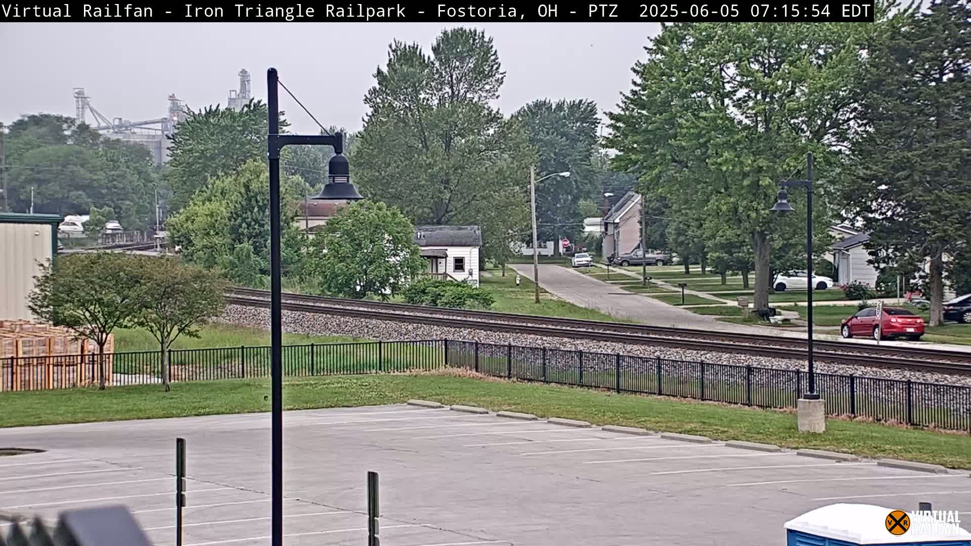A mostly overcast view of a residential street and railroad tracks, with an industrial facility in the distance.