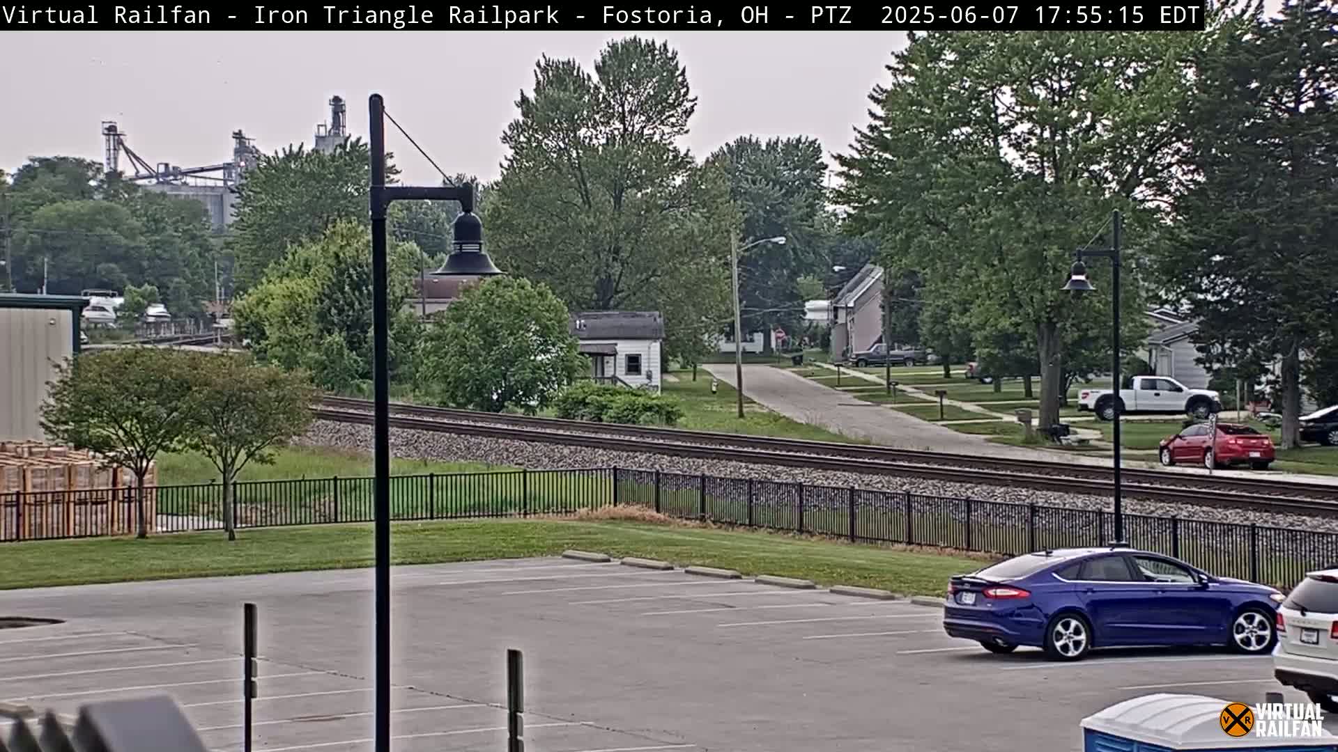 A parking lot overlooks railroad tracks and a residential street, with a factory in the background under an overcast sky.