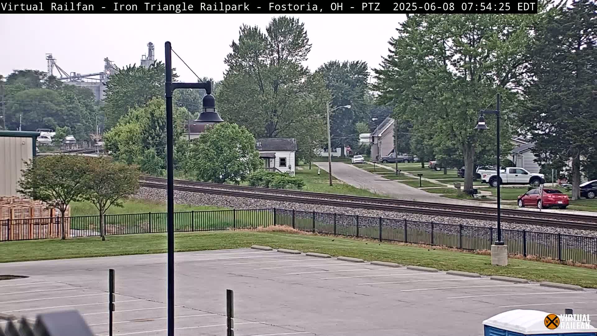 A view of railroad tracks beside a parking lot, with a residential street and industrial structures in the distance on an overcast day.