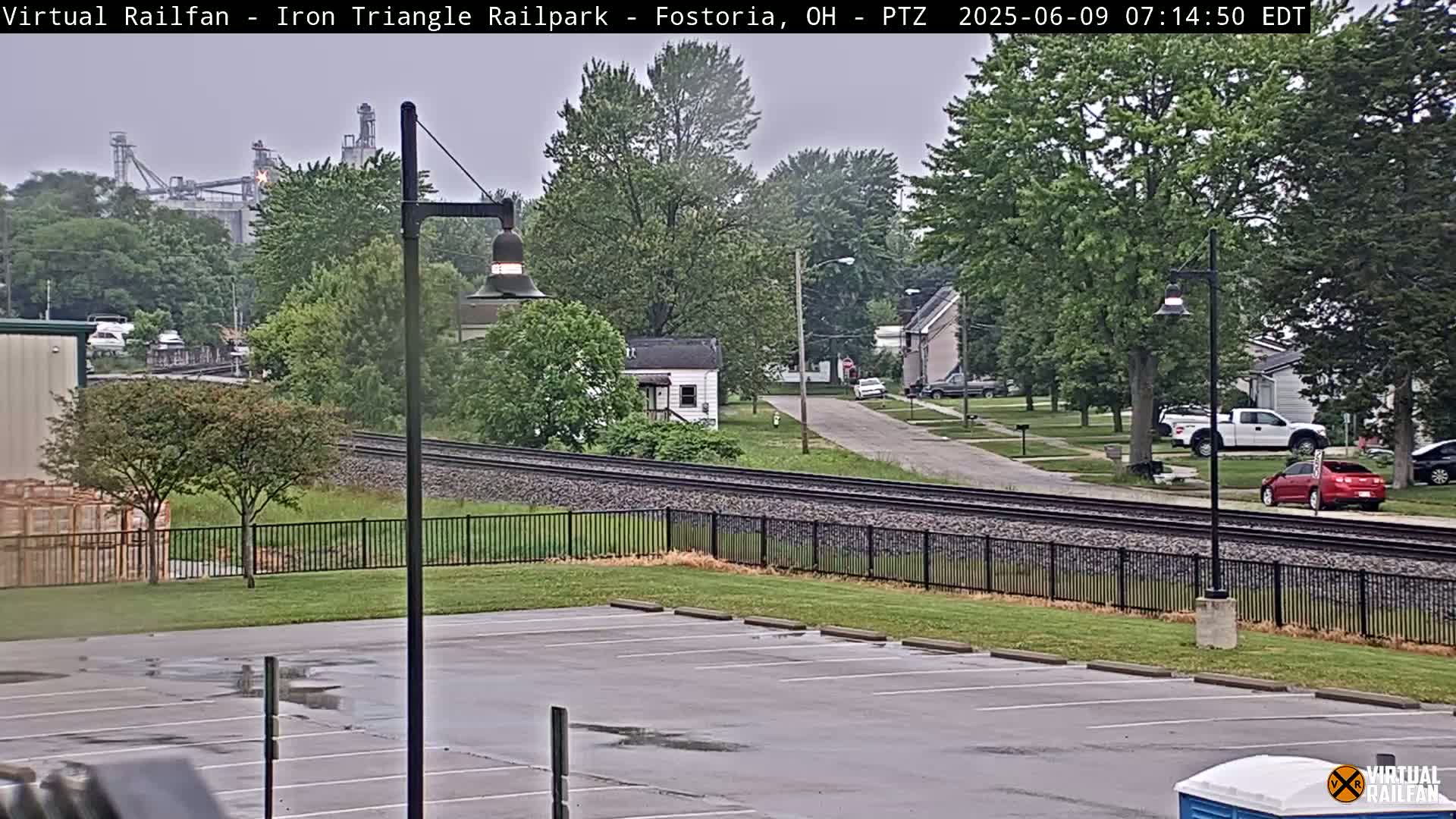 A view of railroad tracks beside a residential street on an overcast day, with industrial buildings visible in the distance.