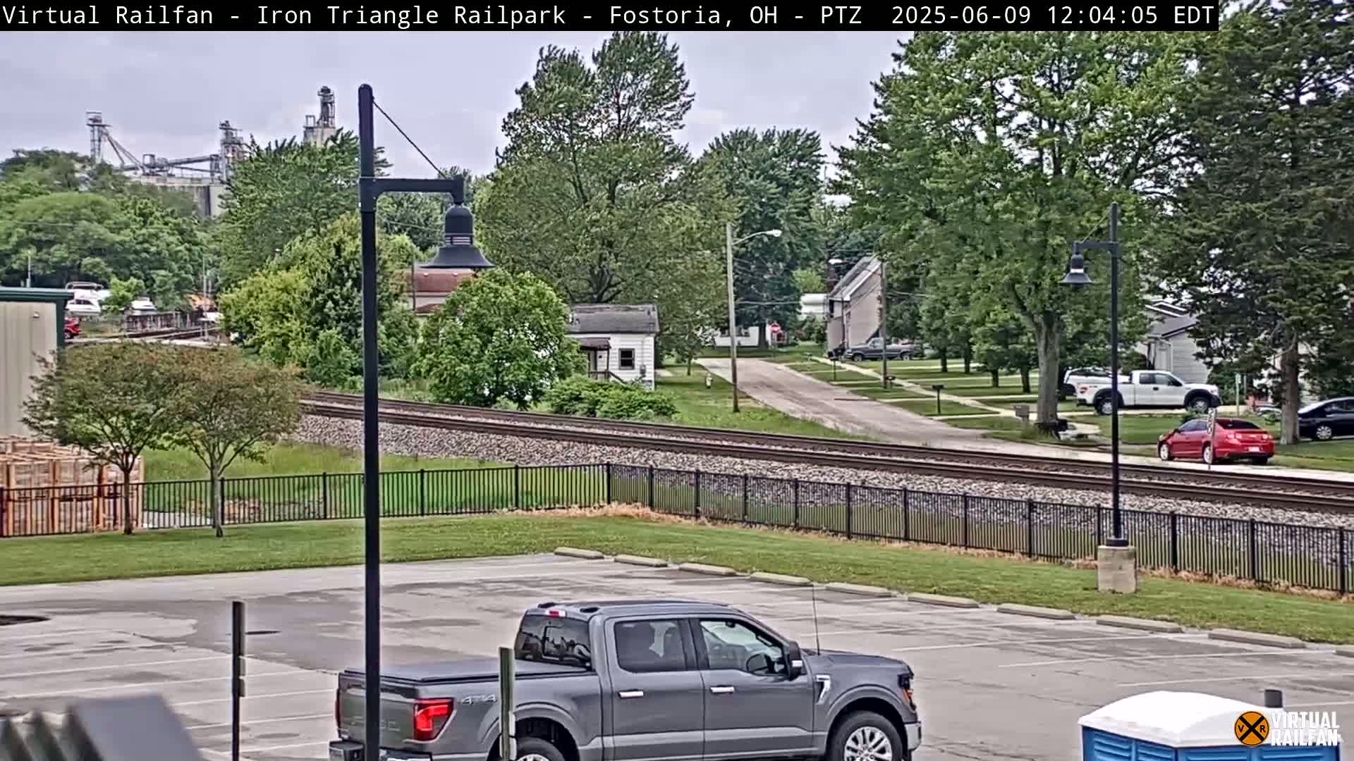A gray pickup truck is parked in a lot next to railroad tracks that run past a residential area on an overcast day.