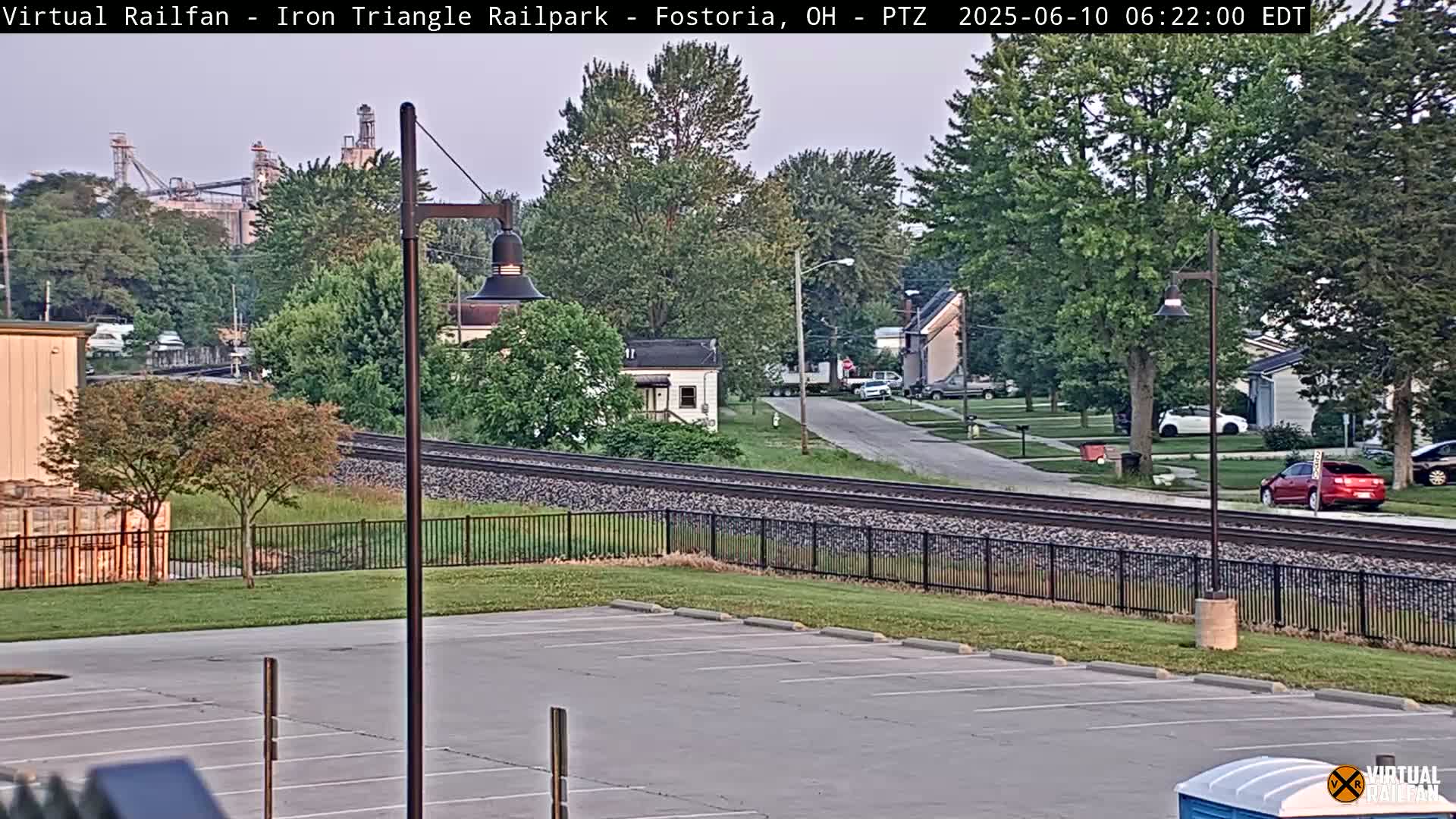 A mostly clear view of a residential street with railroad tracks and a large industrial facility in the background.