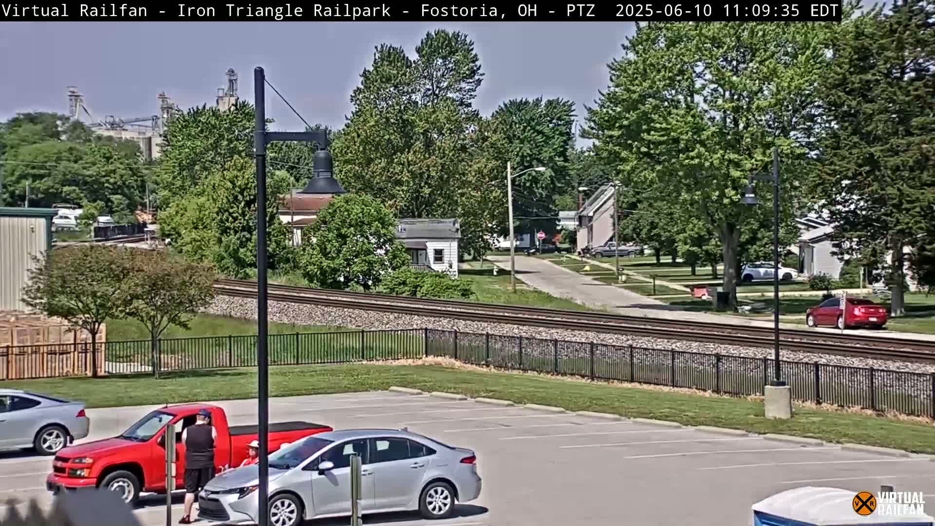 A sunny day shows a parking lot with several cars, including a red pickup truck and a silver sedan, next to railroad tracks that run alongside a residential street with houses and trees, and an industrial area in the background.
