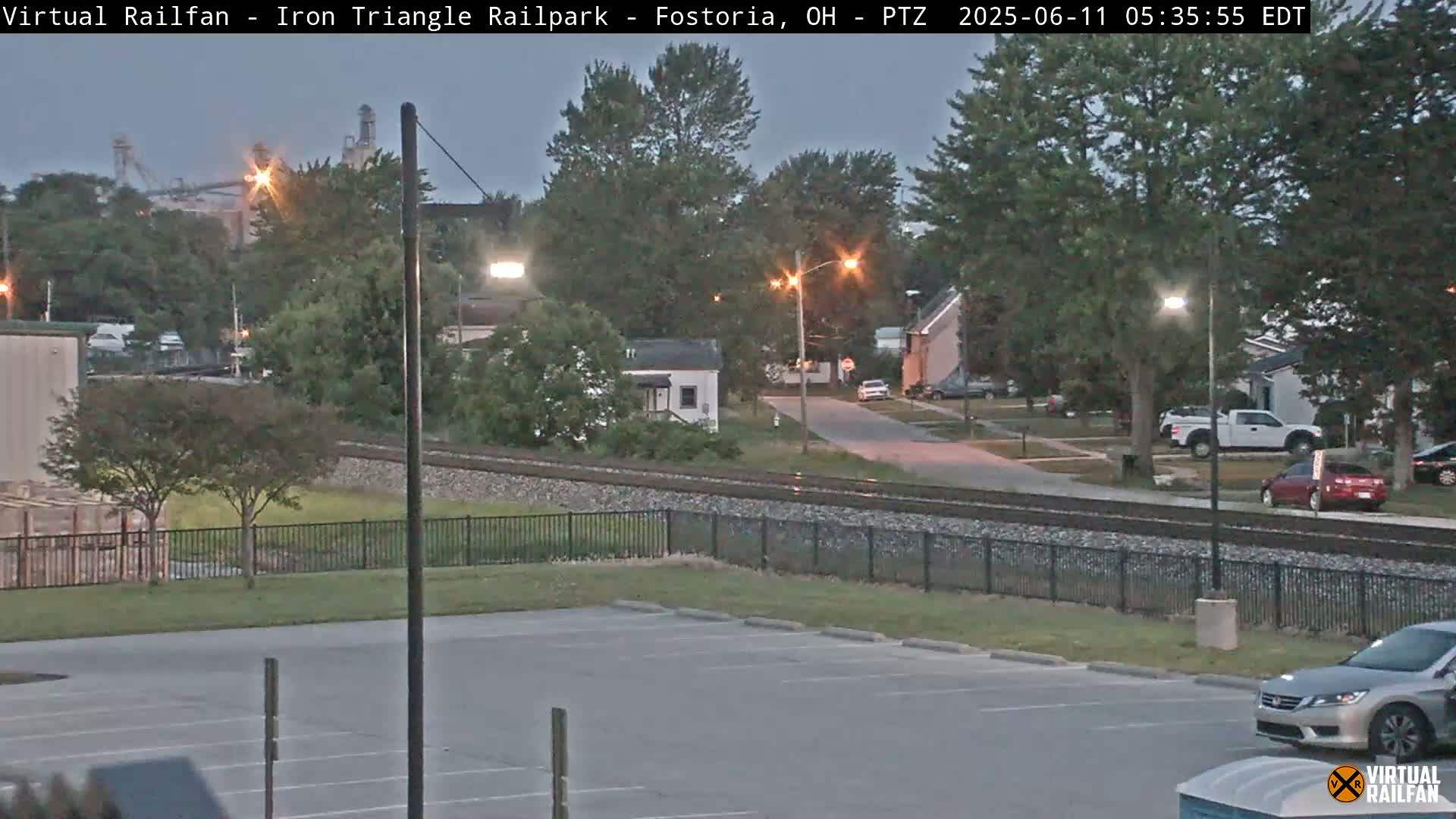 A twilight view of a residential street, railroad tracks, and a parking lot, with a few cars visible and some industrial buildings in the distance.