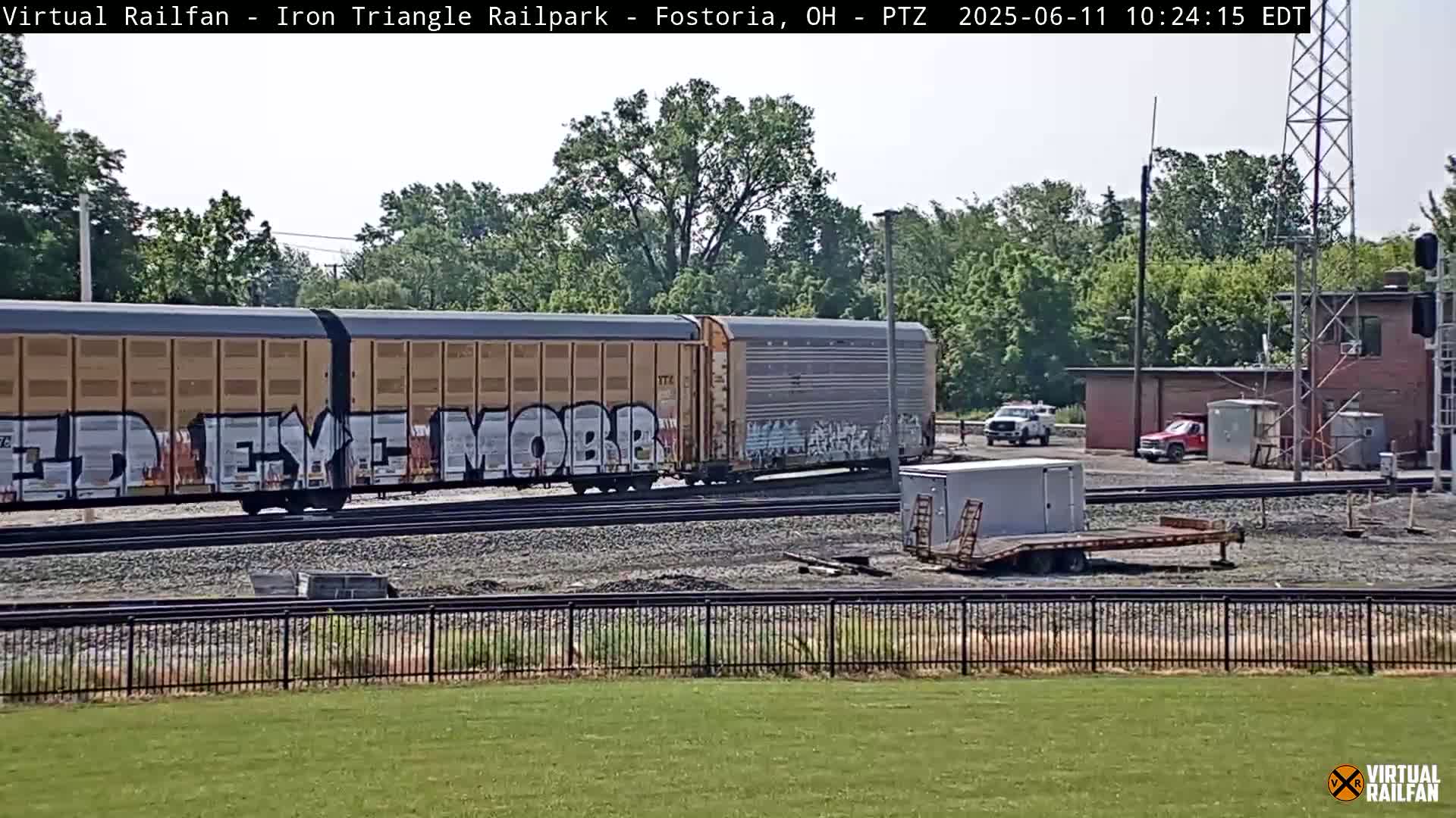 A long freight train with graffiti on its cars sits on railway tracks next to a grassy area, under a partly cloudy sky.