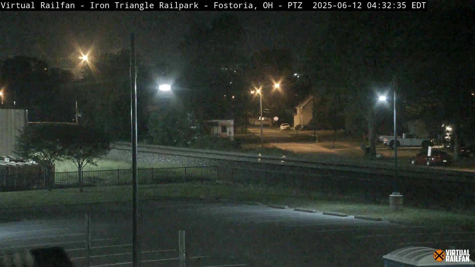 A nighttime view of a railroad track next to a mostly empty parking lot, with several streetlights illuminating the surrounding area.