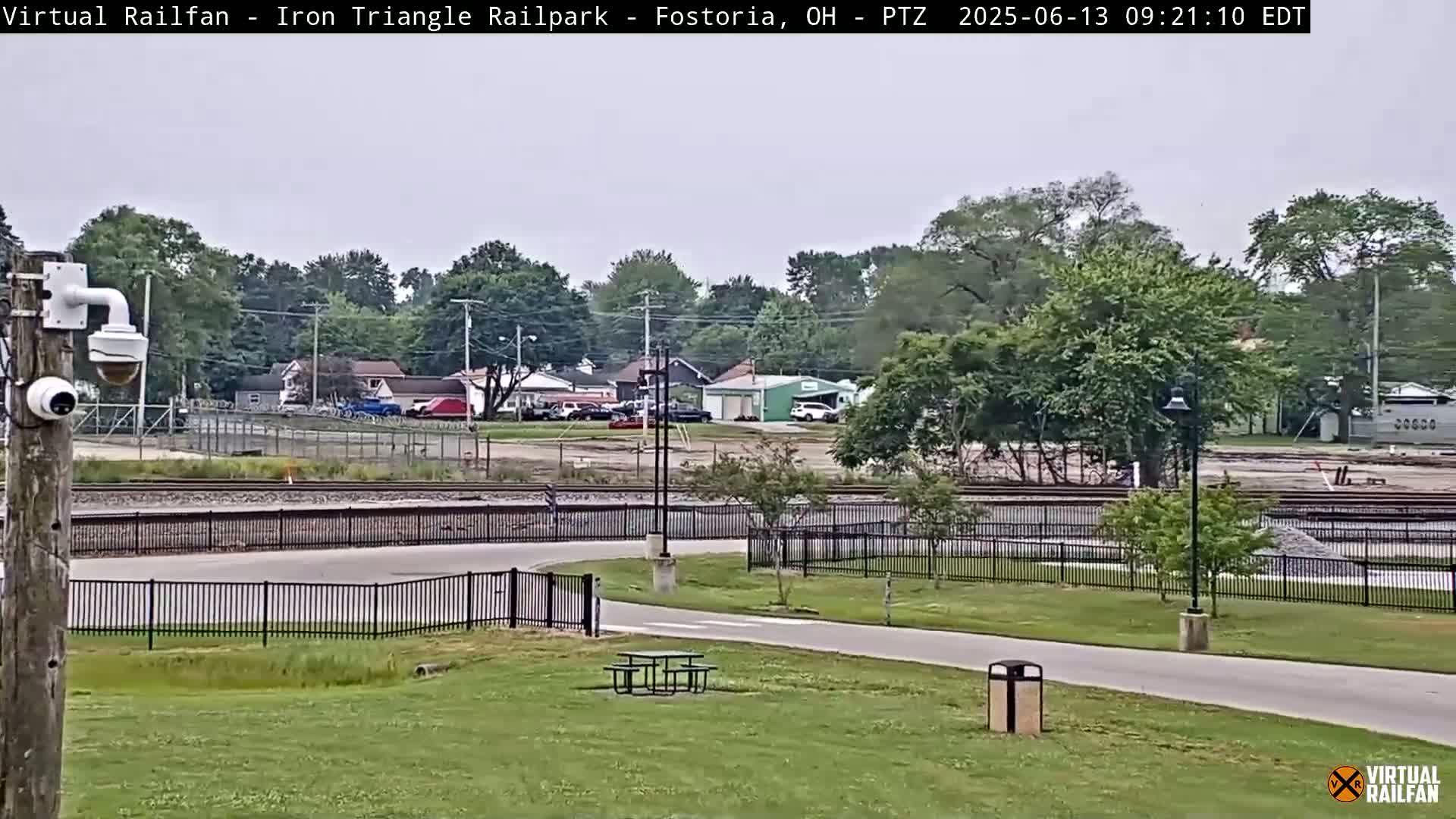 A security camera overlooks a park with a picnic table and a paved path next to railroad tracks and a fenced-in area under an overcast sky.