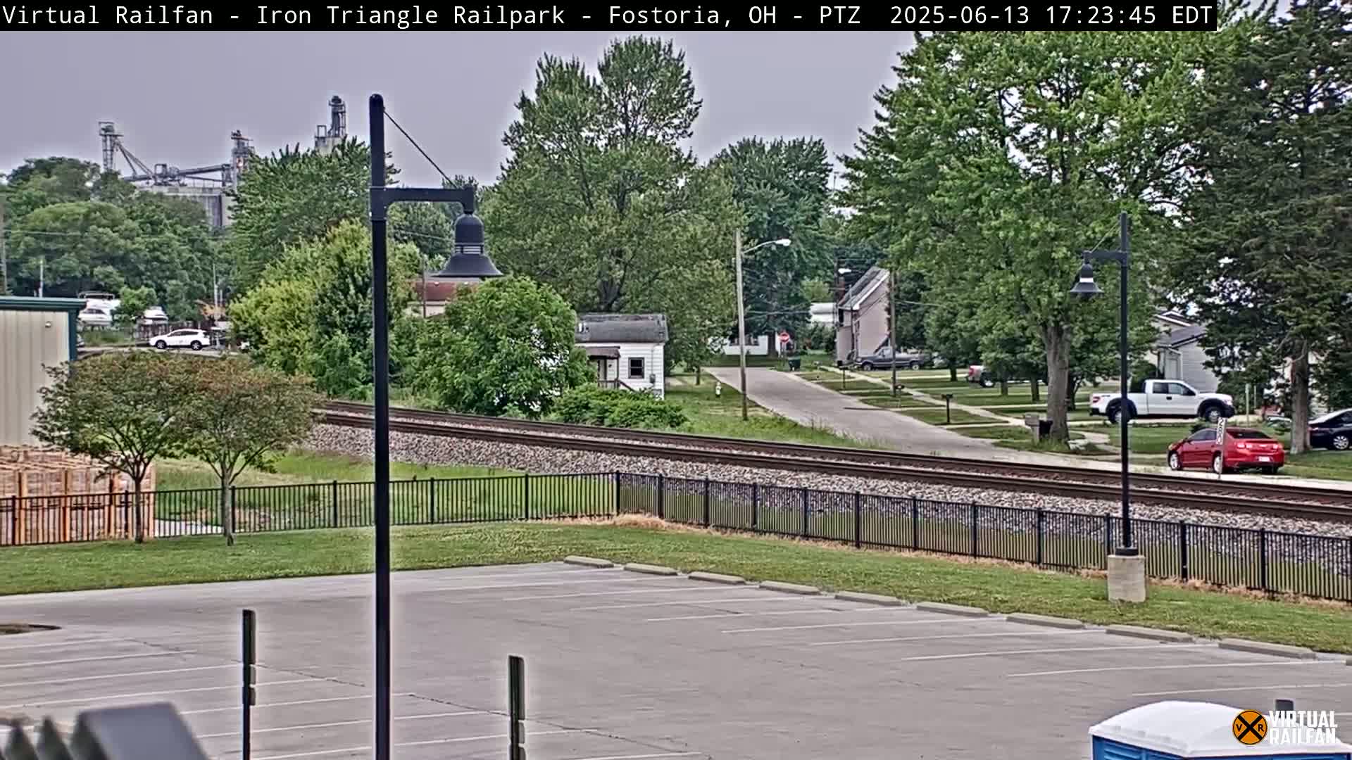 A mostly sunny view of a residential street, railroad tracks, and an empty parking lot, with a factory visible in the background.