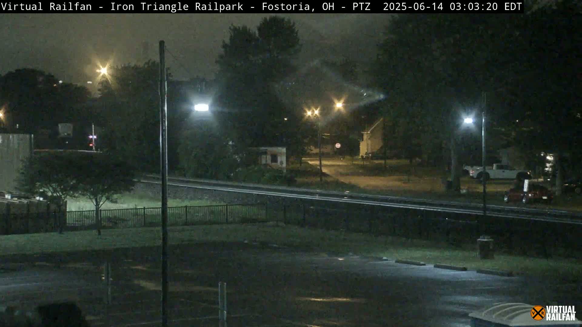 A nighttime view of a rain-slicked parking lot and railroad tracks, with streetlights illuminating nearby trees and a few parked cars.