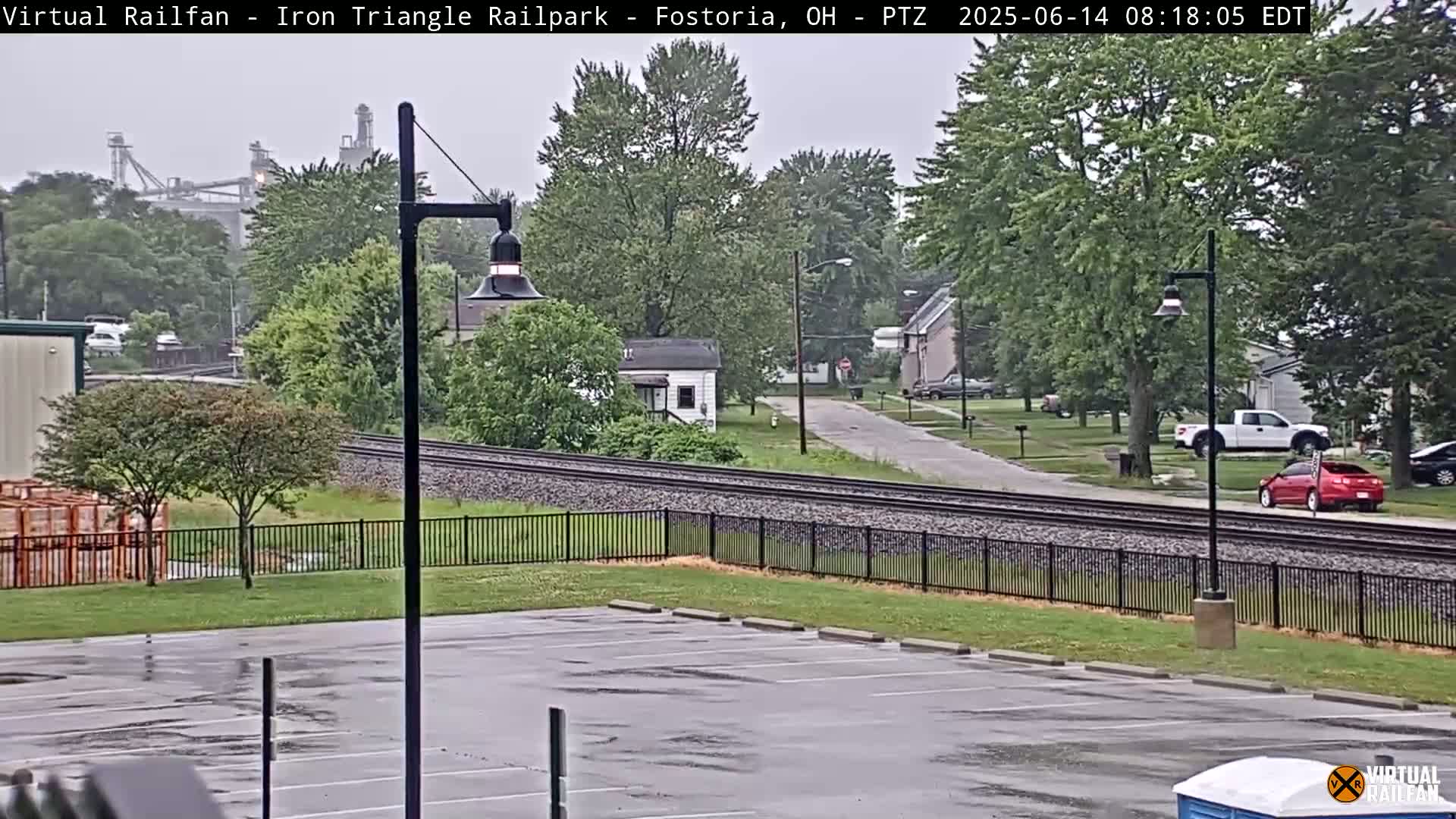 A wet parking lot overlooks railroad tracks and a residential street with houses and a distant industrial area under an overcast sky.