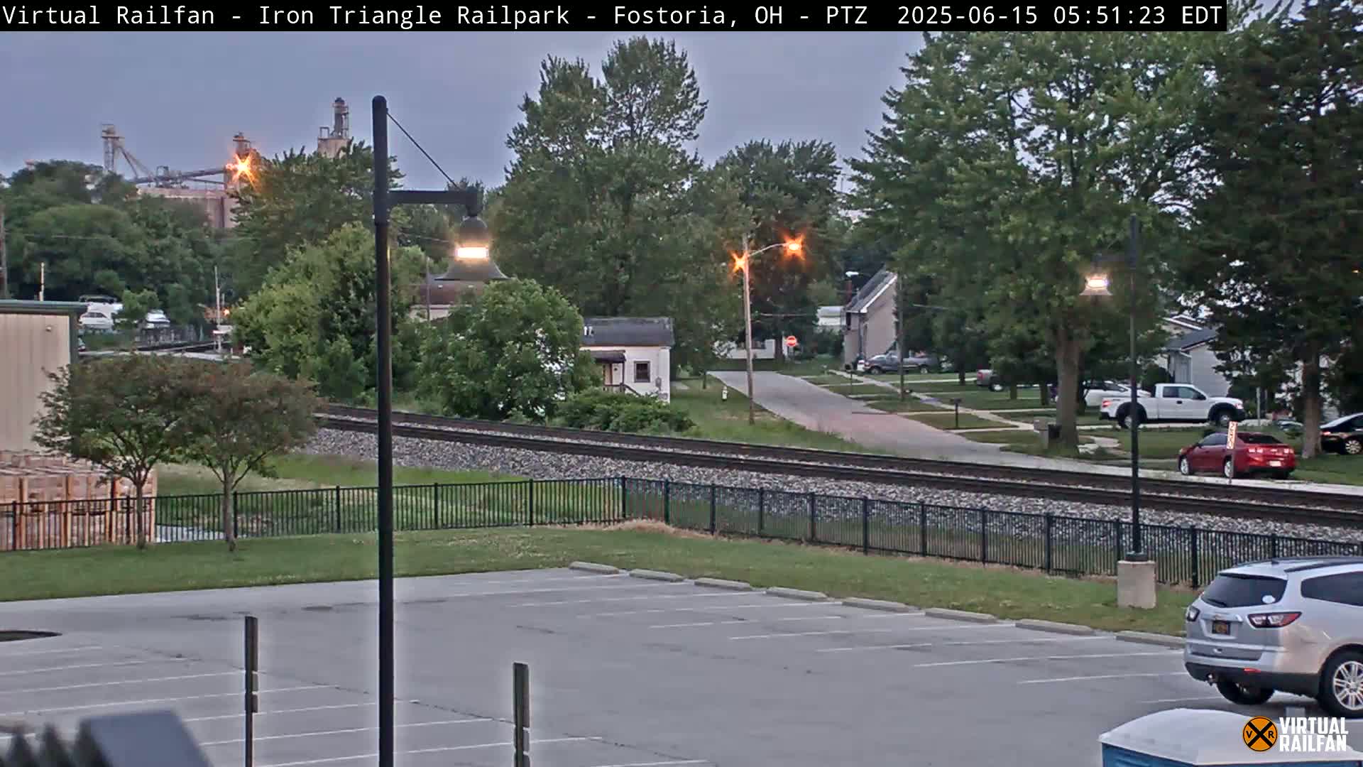 A mostly empty parking lot overlooks railroad tracks that run through a residential area under a twilight sky.
