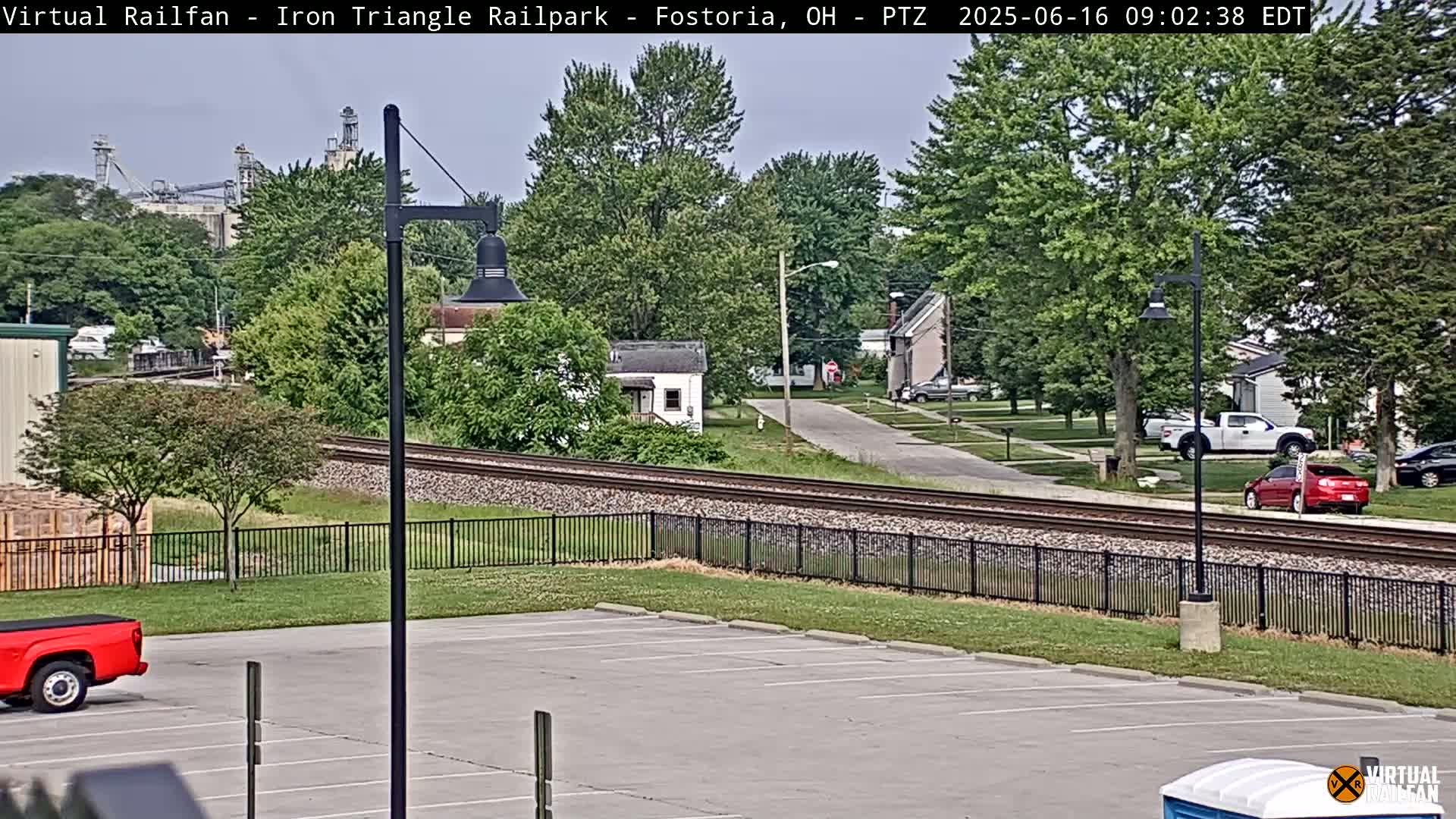 A mostly sunny day shows a view of railroad tracks beside a residential area with parked cars, a parking lot, and trees.