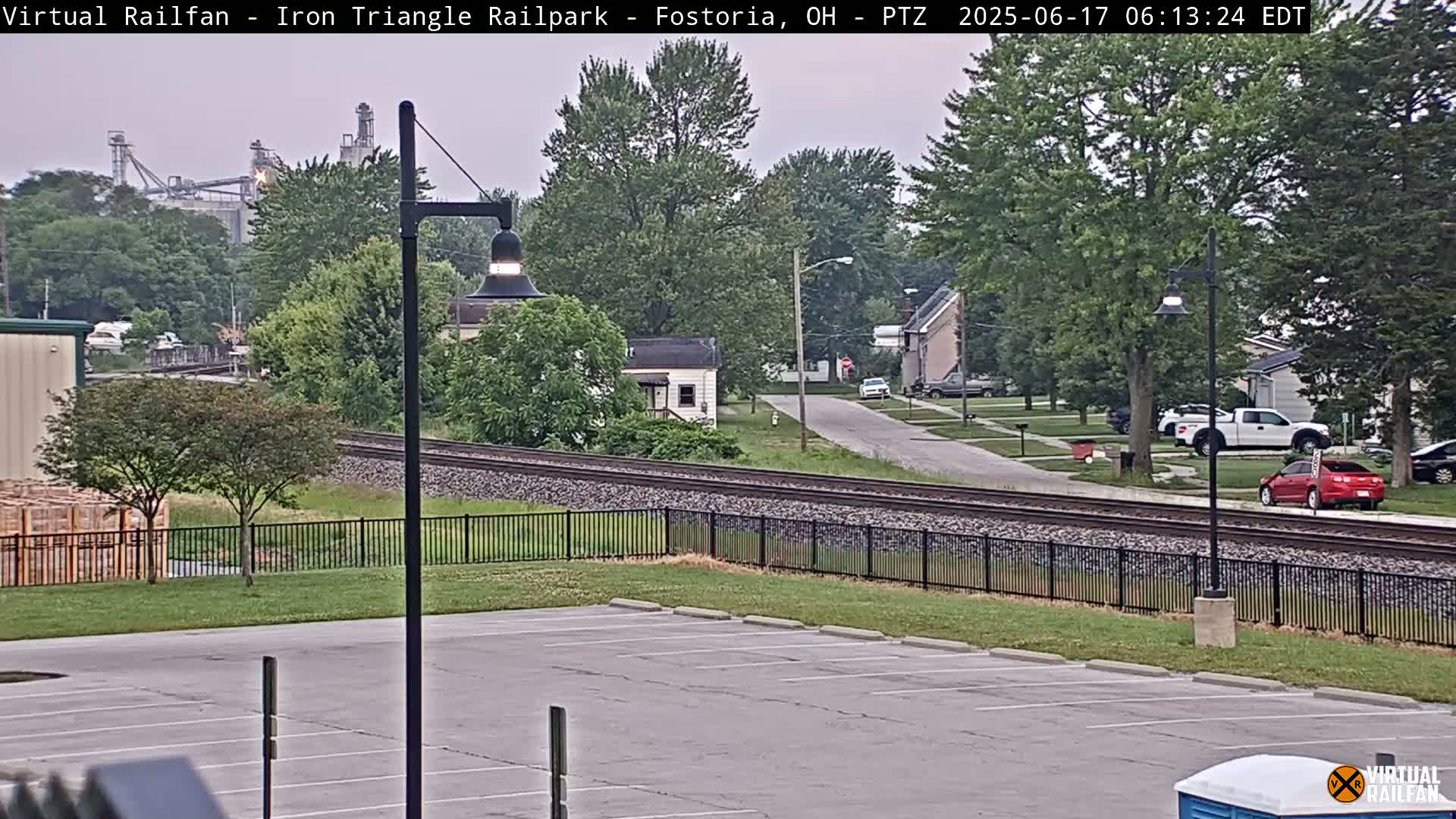 A mostly cloudy, overcast view of a residential street with houses, a parking lot, railroad tracks, and an industrial facility in the background.