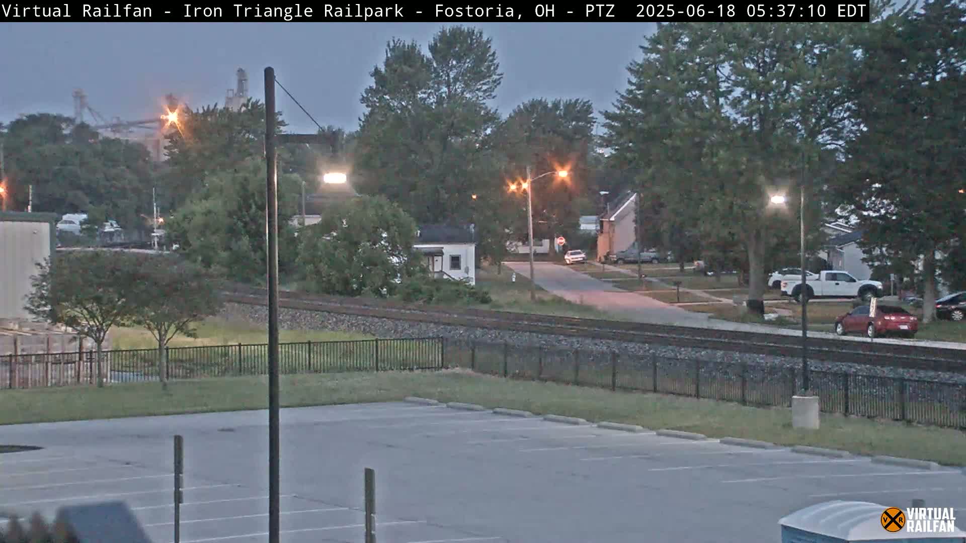 A twilight view of an empty parking lot next to railroad tracks, beyond which are residential streets lined with trees and houses under a dusky sky.