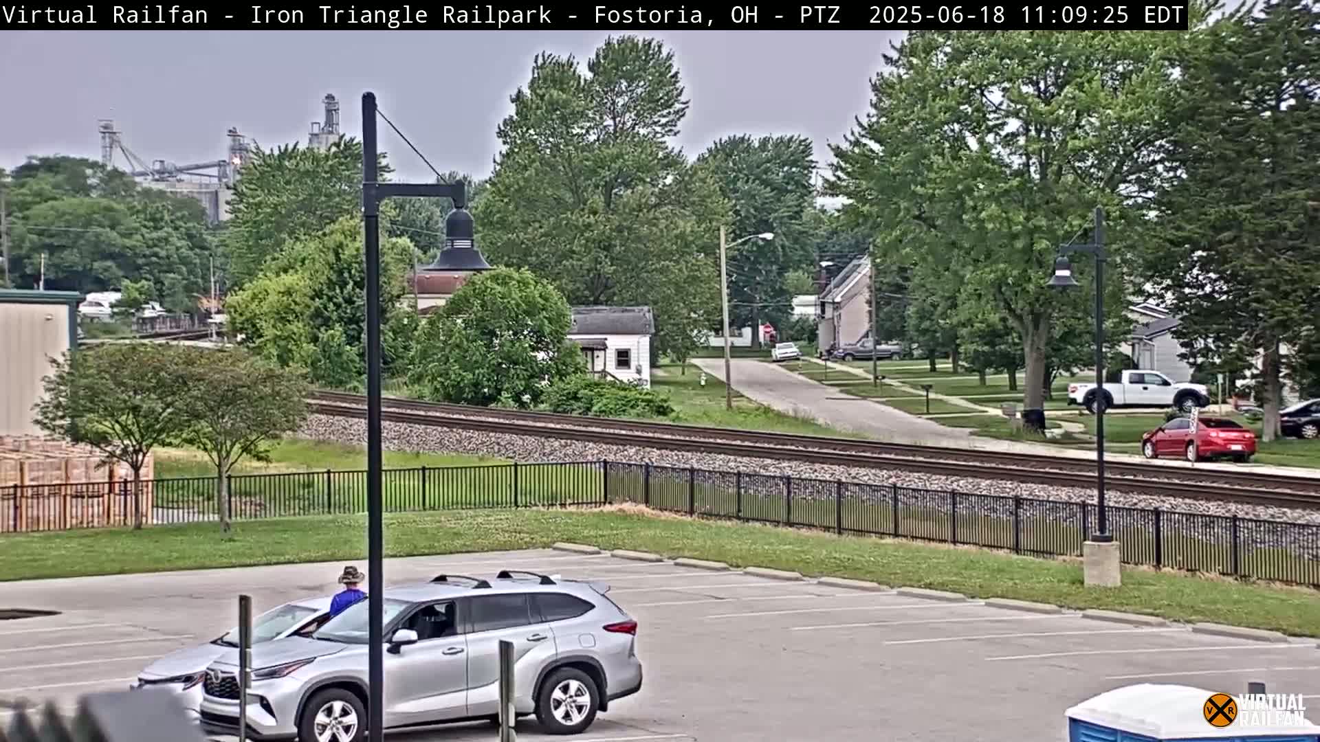 A silver SUV is parked in a lot next to a railroad track, with a person in a blue shirt and hat sitting inside, under an overcast sky.