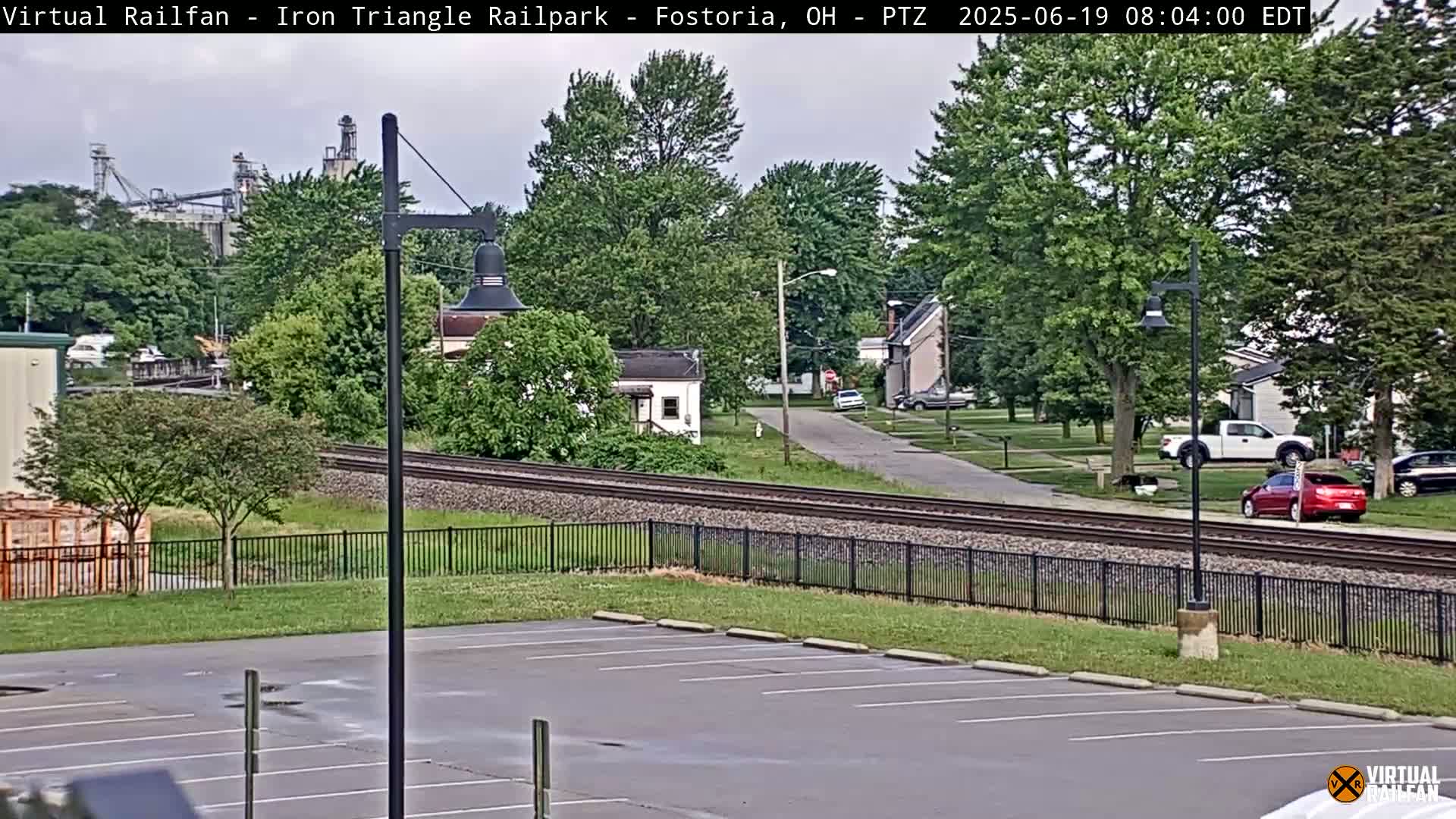 A mostly cloudy day shows a residential street crossing railroad tracks, with a parking lot and industrial buildings in the background.