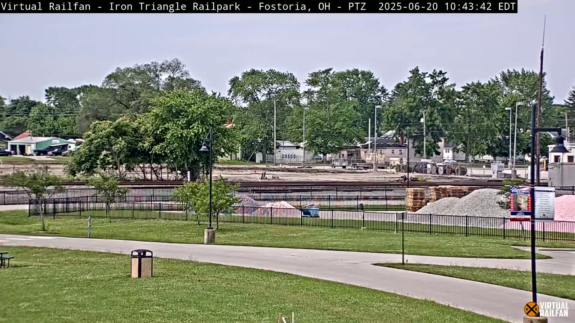 A mostly sunny day reveals a park with a paved path, green grass, and a construction area behind a black fence, featuring piles of gravel and railroad tracks.