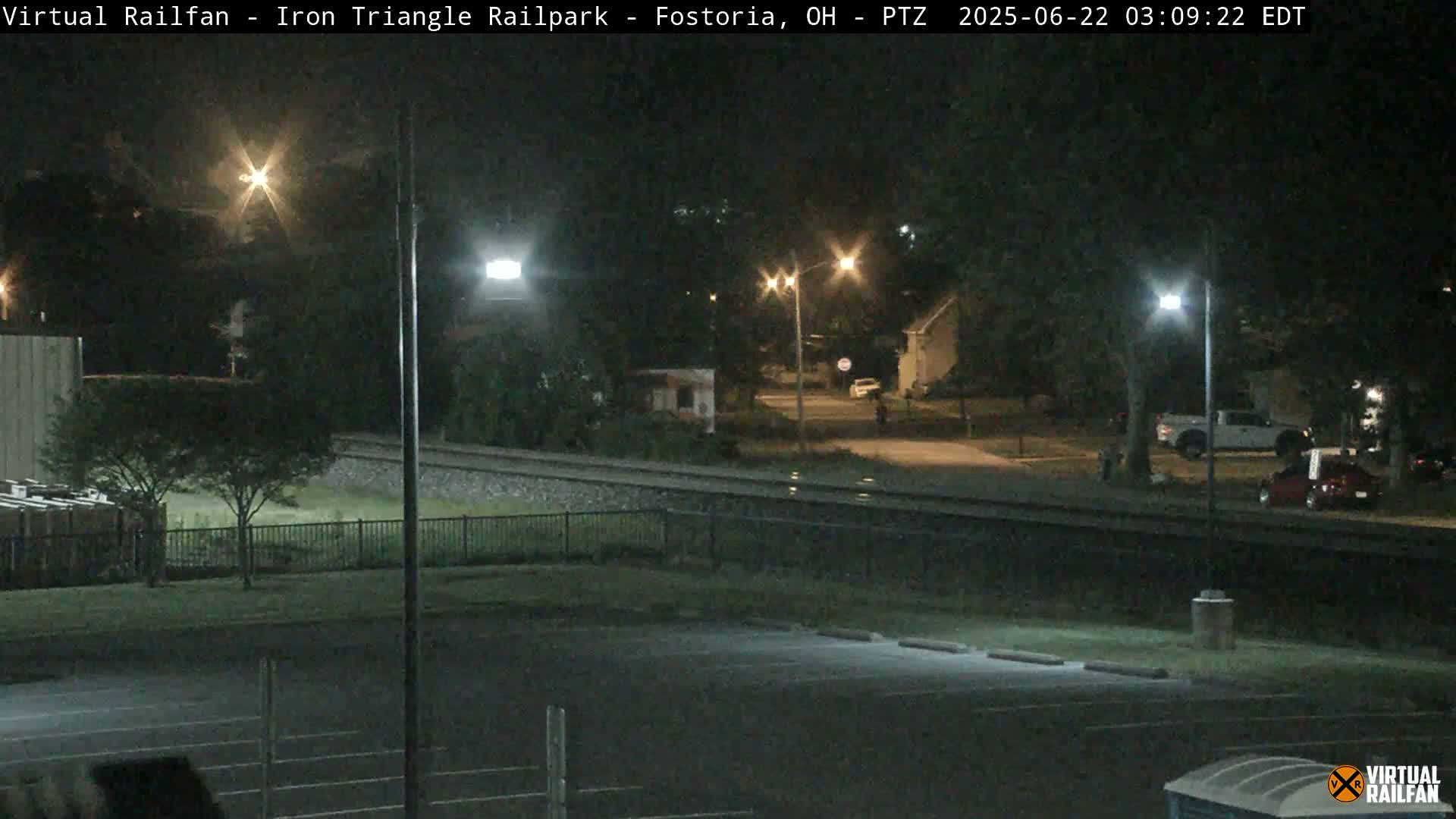 A nighttime view of a railroad track next to a parking lot and residential street, illuminated by streetlights.