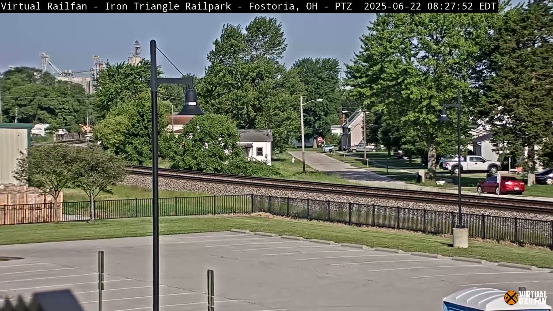 A mostly sunny day shows a view of railroad tracks next to a residential area, with an empty parking lot in the foreground.