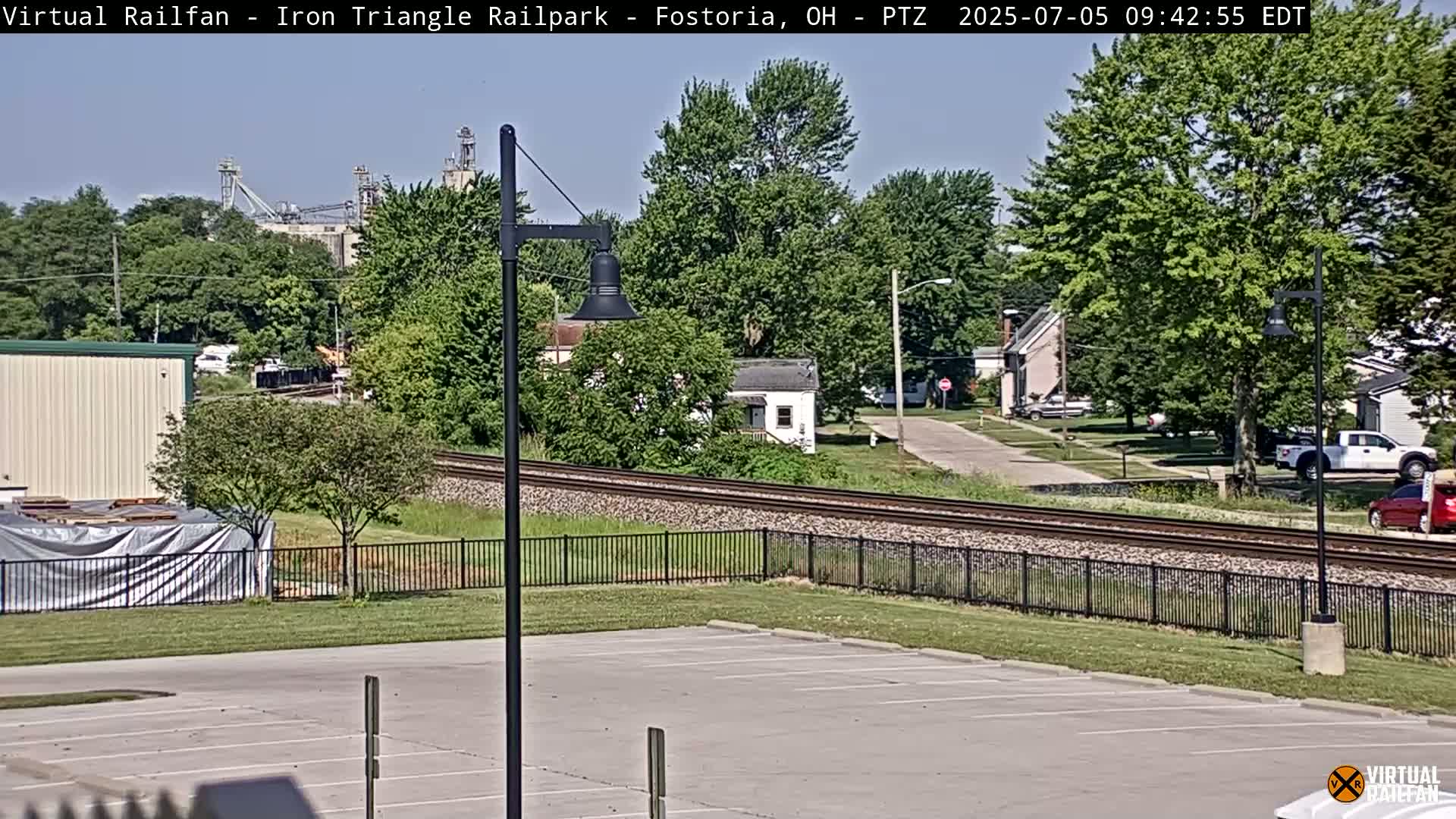 A mostly sunny view of a parking lot next to railroad tracks, with a residential area and industrial buildings in the background.