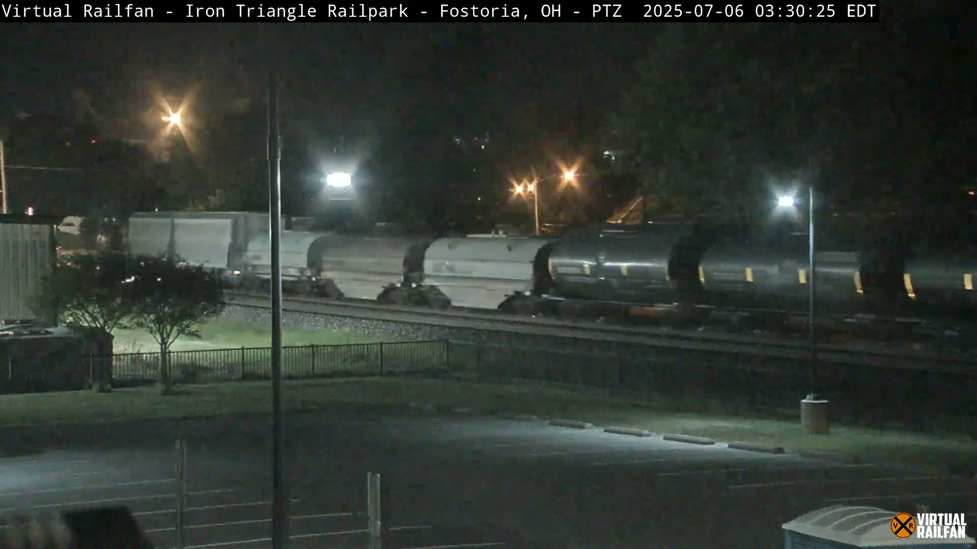 A long freight train of tanker cars moves through a rail yard at night.