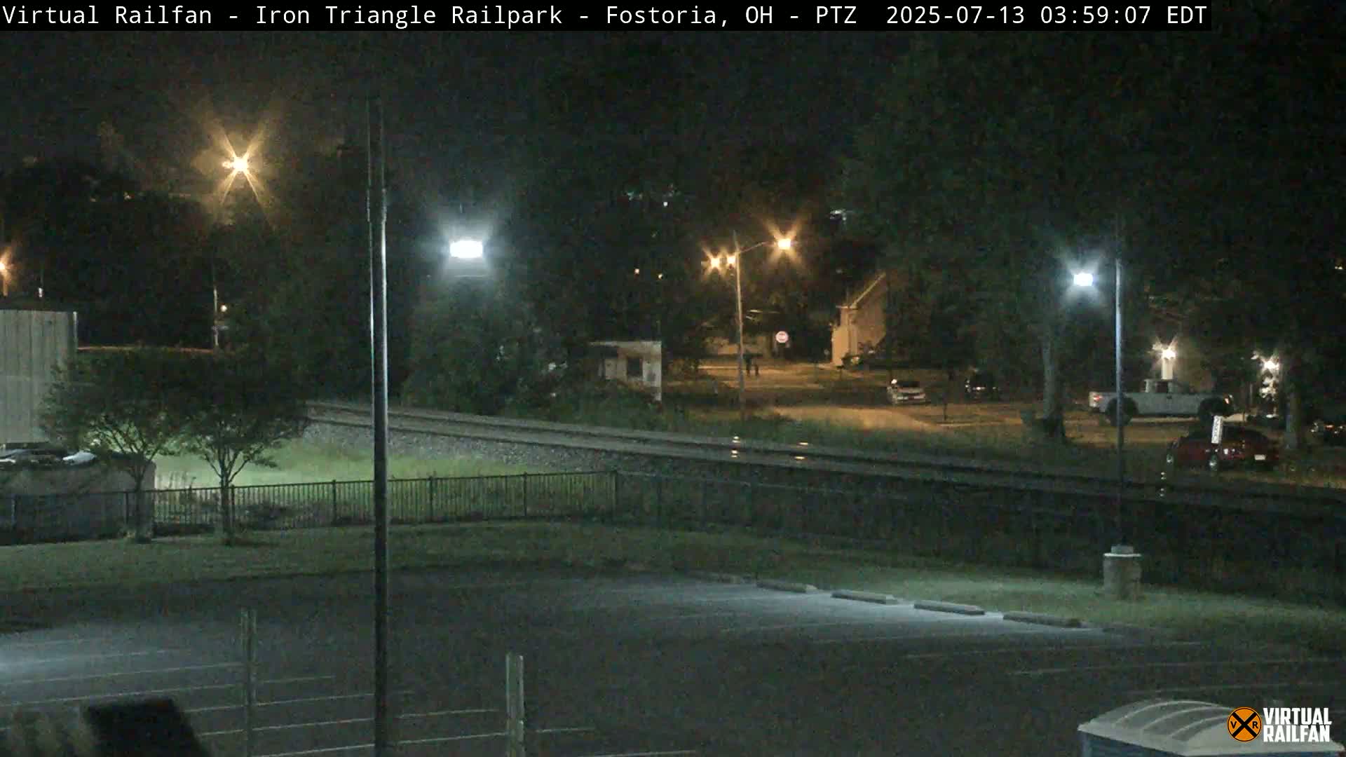 A nighttime view of a rail yard and adjacent street, illuminated by streetlights, with several vehicles parked nearby.