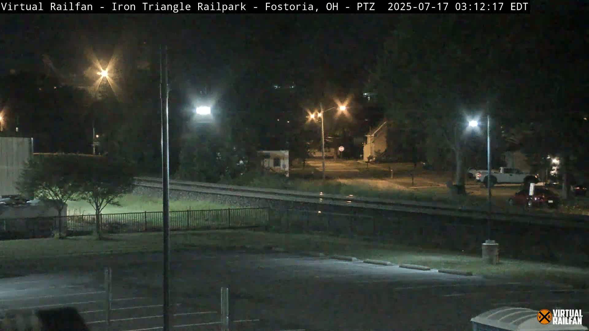 A nighttime view of a railroad track next to a parking lot and residential area, illuminated by streetlights.