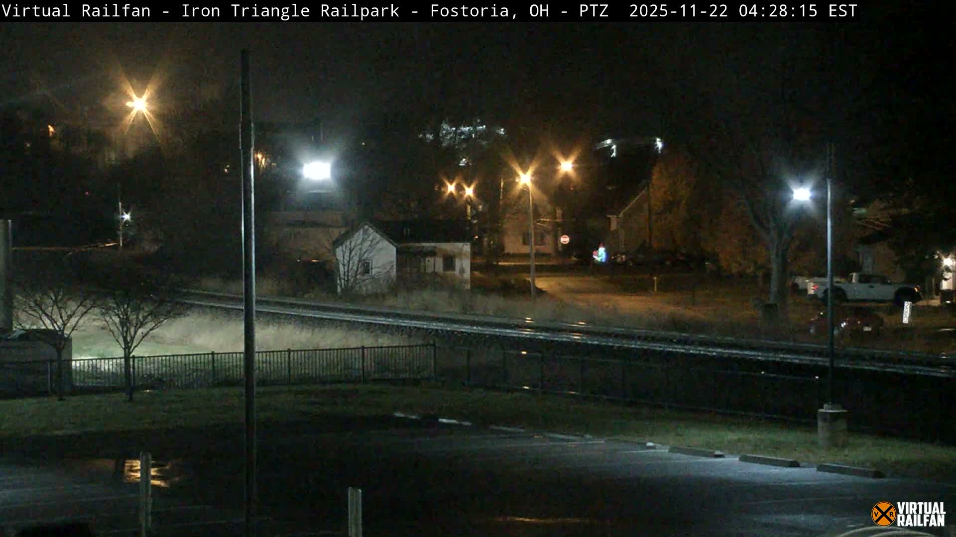 The image depicts a damp night scene of railroad tracks with a bright train headlight approaching on the left, bordered by streetlights, buildings, and bare trees, with a wet parking lot visible in the foreground.