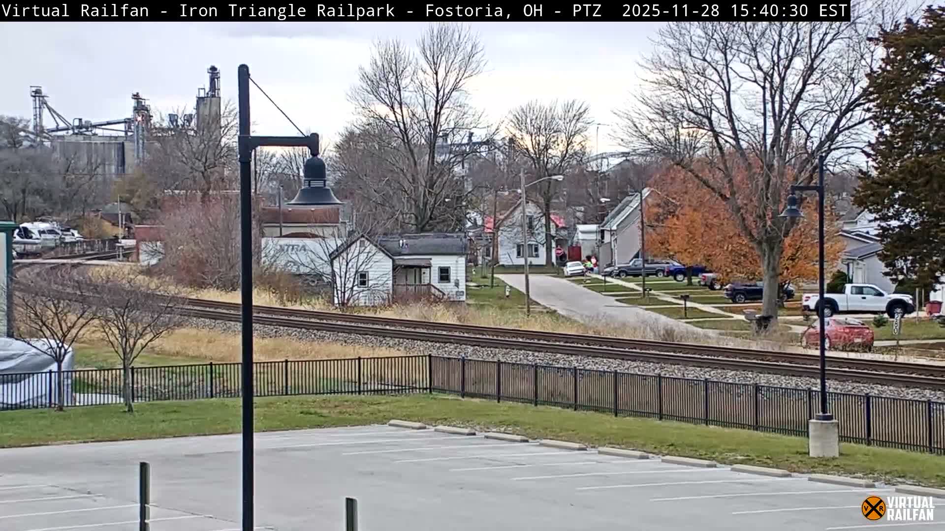 A cloudy, late autumn day reveals a townscape featuring a double railroad track running horizontally across the mid-ground, surrounded by residential houses, a mix of bare and orange-leafed trees, and an industrial complex in the background.