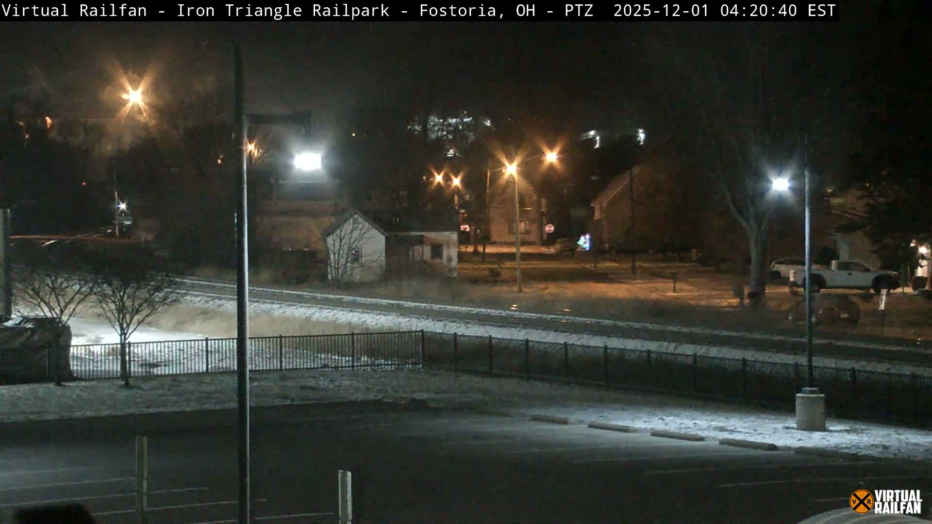 A quiet, clear winter night scene shows a light dusting of snow covering railroad tracks, a parking lot, and ground, with streetlights illuminating nearby houses, bare trees, and a bright, distant light.