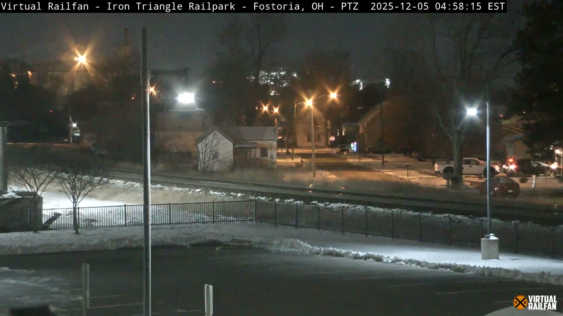 A snowy winter night scene reveals railroad tracks running between snow-dusted ground, illuminated streets, houses, and distant vehicle lights.