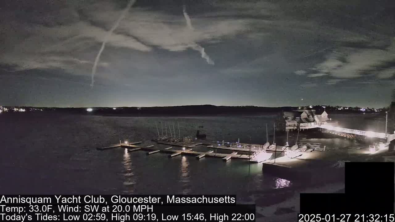A nighttime view of a yacht club harbor with boats docked, under a cloudy sky with a contrail visible.