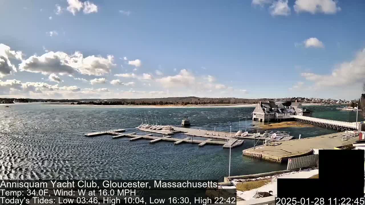 A marina with numerous boats docked at slips, a clubhouse, and a long pier extends into a choppy body of water under a partly sunny sky.