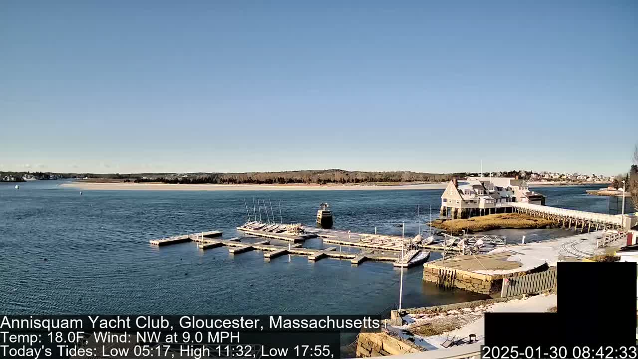 A marina with docked sailboats is situated on a calm body of water under a clear blue sky, with a shoreline and distant land visible in the background, and a light dusting of snow on the ground.