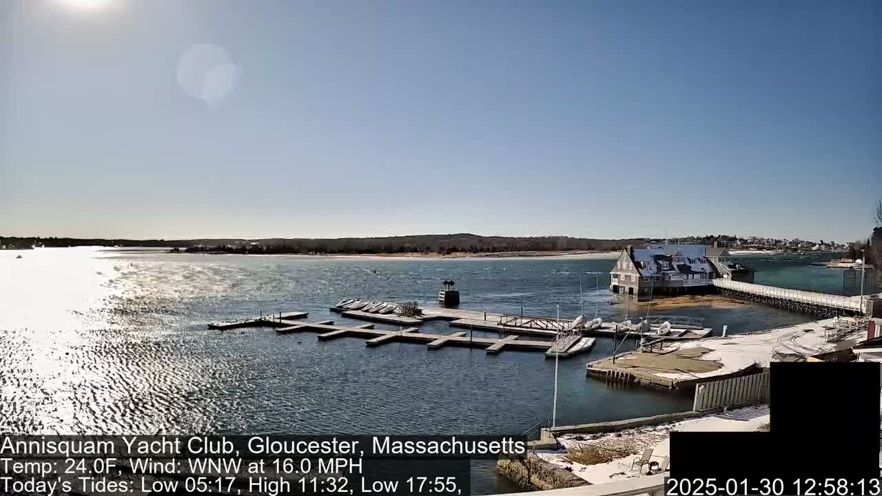 A sunny day shows a harbor with docked sailboats, a clubhouse, and a long dock extending into the water, under a mostly clear sky with a light chop on the water.