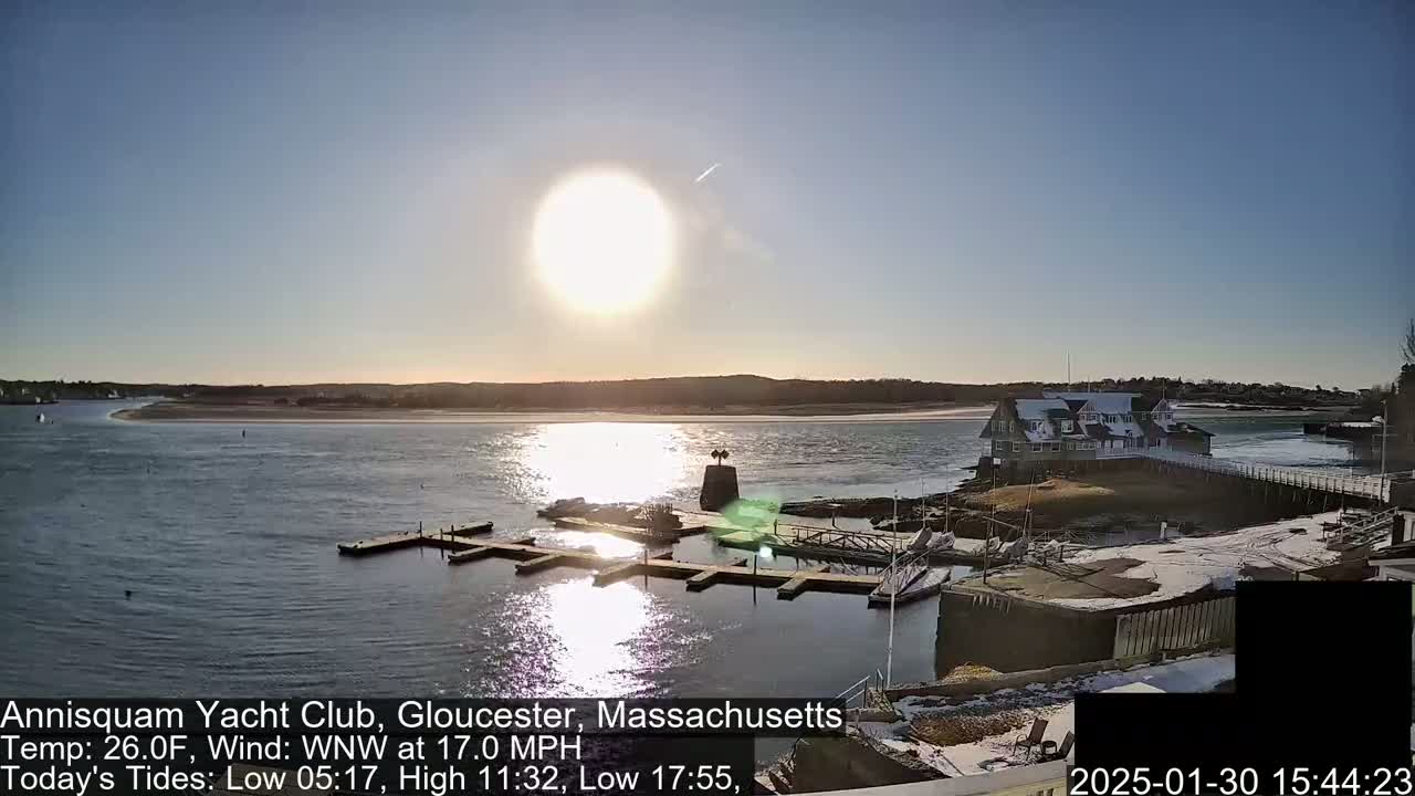 A sunny day at a marina with a calm body of water, docks, a building, and some snow on the ground.