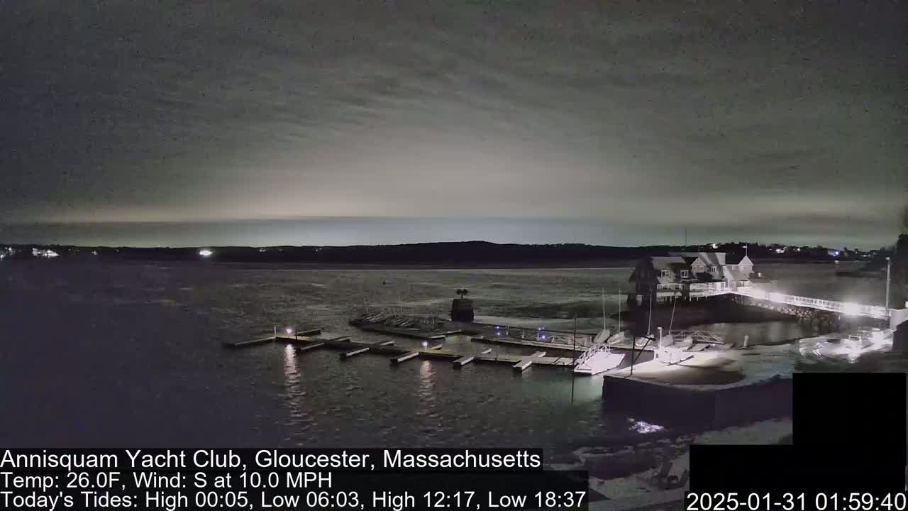 A nighttime view of a marina with docked boats, a clubhouse, and a calm body of water under a cloudy sky.