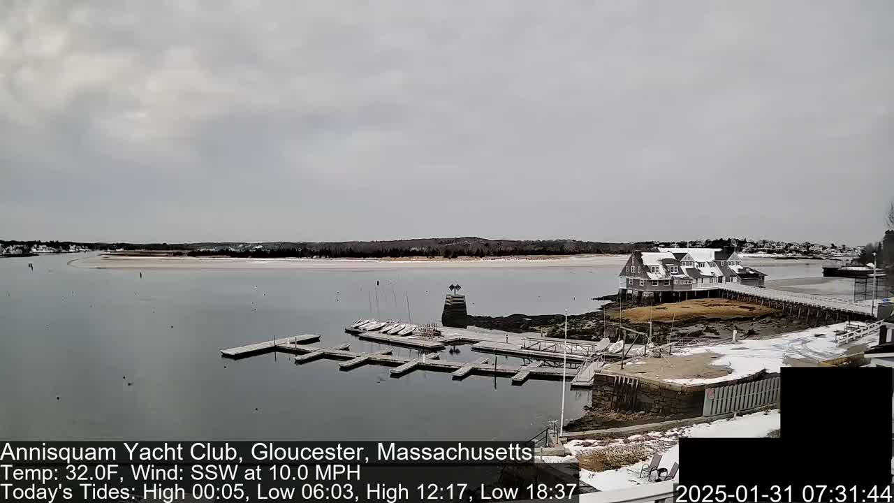 A marina with docked boats and a clubhouse is situated on a calm body of water under an overcast sky; there is snow on the ground.