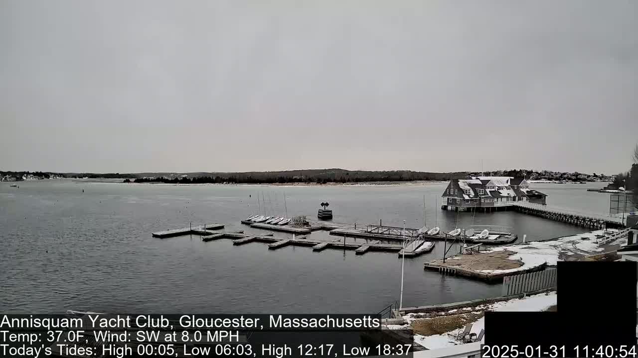 A calm, overcast day shows a marina with numerous small sailboats docked at a waterfront clubhouse, with some snow visible on the ground and nearby structures.